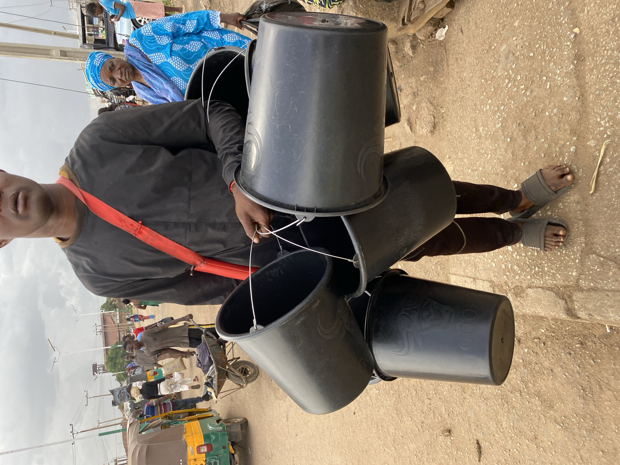 A person is carrying several black plastic buckets in an outdoor market setting in Bukuru, Nigeria. The buckets appear to be stacked and are attached by their wire handles. The person is wearing a black shirt and dark pants, with an orange strap across their chest. Their feet are visible, wearing gray flip-flops.
In the background, other people are seen, including women in brightly colored headscarves and traditional clothing. Some are standing near a vehicle, possibly a three-wheeled auto-rickshaw, and a cart. The ground is dusty and appears to be unpaved. The sky is overcast, suggesting cloudy weather. The scene depicts a busy market environment, with individuals engaged in daily activities. The overall impression is one of a typical day in a Nigerian market. No visible text is present in the image.