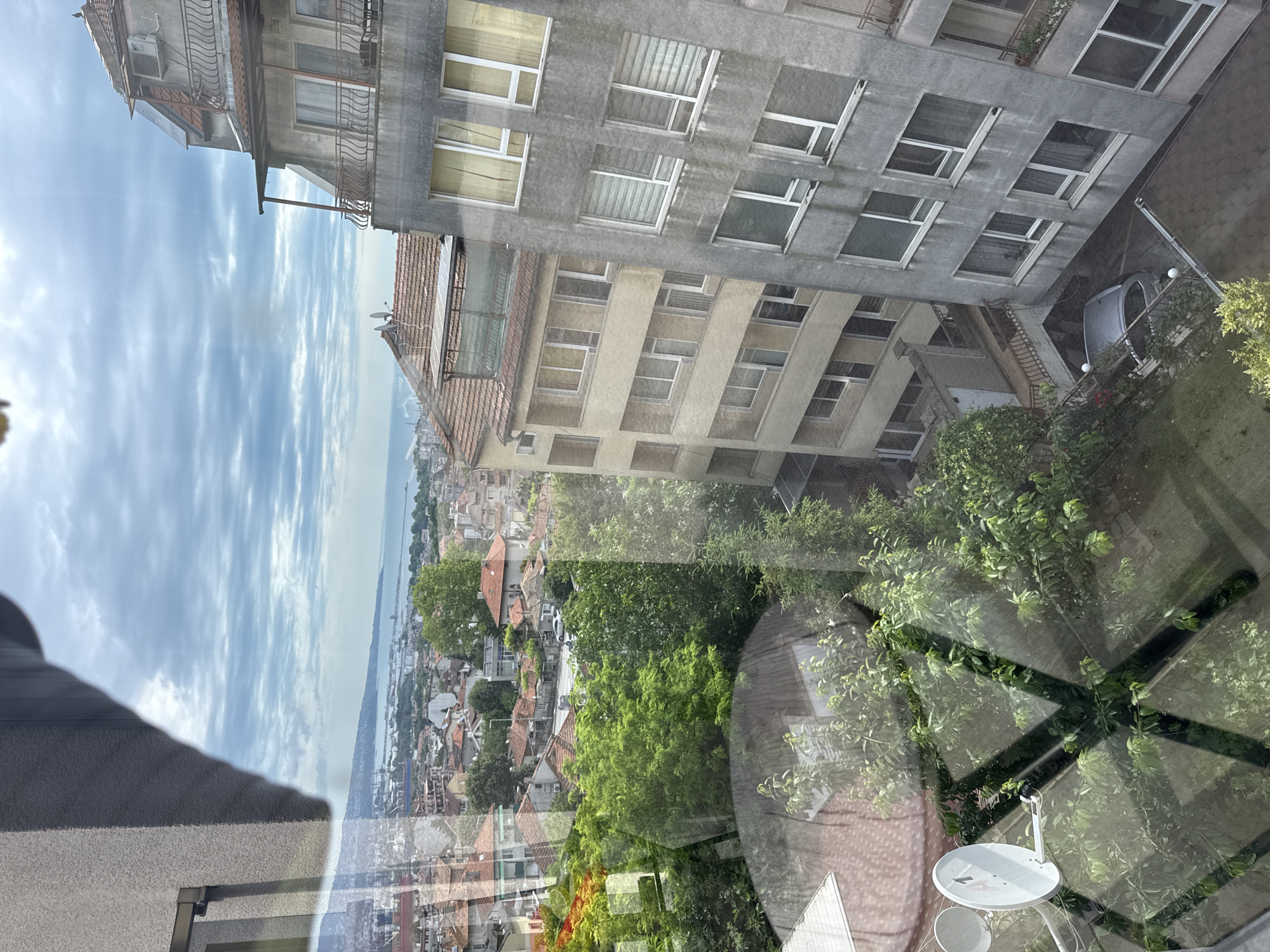 The image shows a view from a window looking out over a city with buildings and a body of water in the distance. In the foreground, a satellite dish is visible, along with reflections on the window. The middle ground consists of several apartment buildings of varying heights and architectural styles, interspersed with lush green trees and red-tiled roofs. The buildings are close together, suggesting an urban environment. In the background, a coastline stretches out towards the horizon, with a hazy sky above, characterized by scattered clouds. A grey car is parked in an open area between two buildings in the lower right portion of the frame. There are no people visible in the image. The overall scene depicts a dense urban landscape with elements of nature and a distant view of the sea.