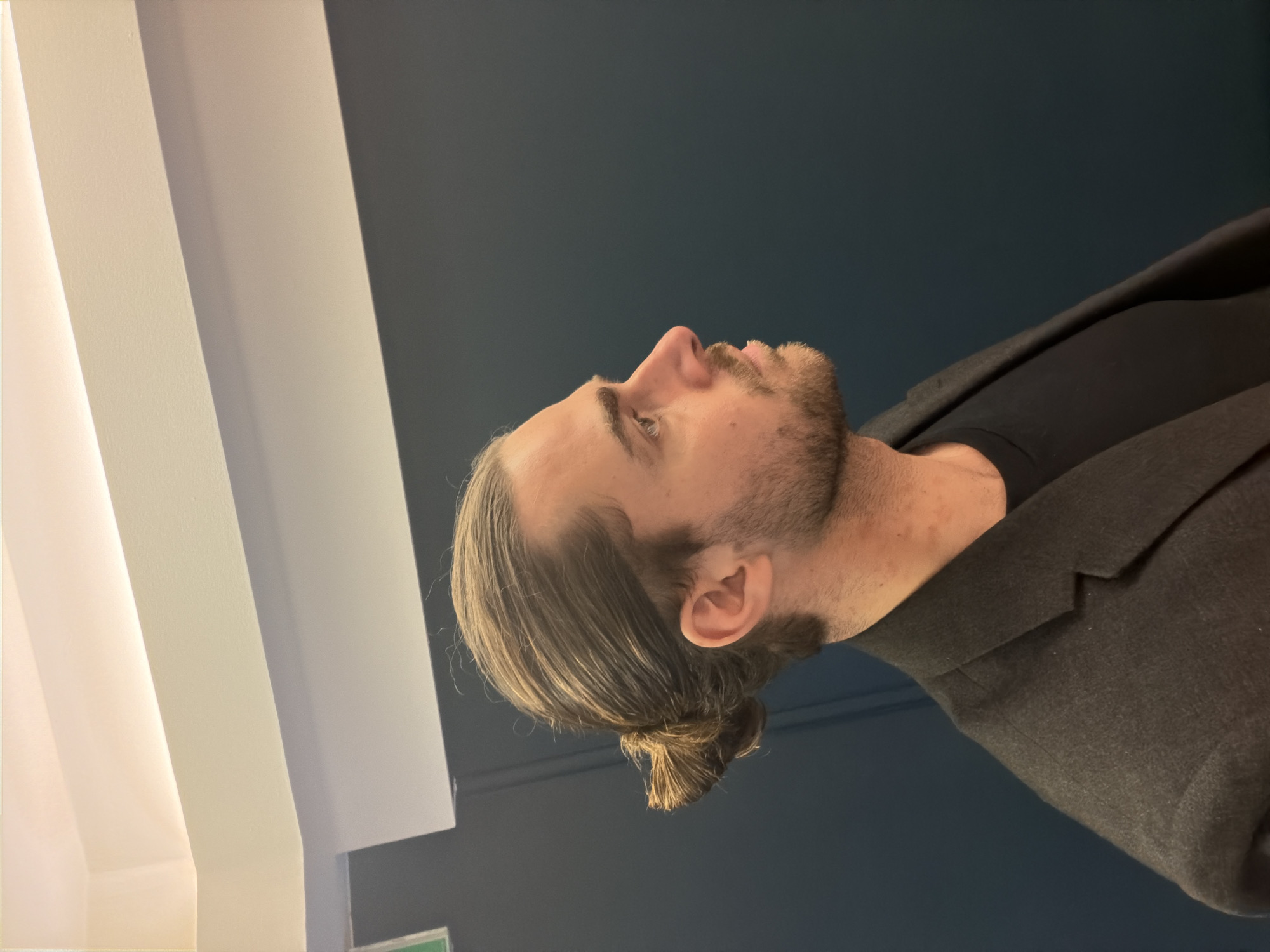 A man with long, slicked-back brown hair tied in a bun and a trimmed beard is looking upwards. He's wearing a dark, possibly black, t-shirt underneath a gray blazer. The background consists of a dark blue wall and what appears to be a white architectural structure with parallel lines. The location cannot be determined to be Paris, France from the image alone.