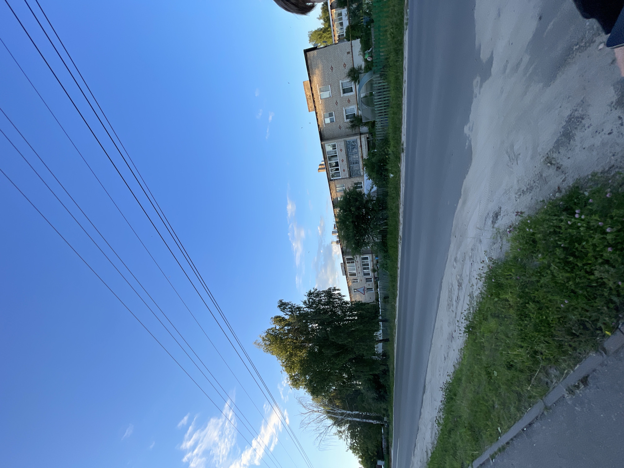 The image shows a residential area on a clear day. In the foreground, there is a paved road with a grassy shoulder. Beyond the road, there is a multi-story apartment building made of light-colored brick. Several trees are located between the road and the building. The sky is blue with a few scattered clouds. Power lines are visible spanning across the upper portion of the image. A partial view of a person's head and shoulder is visible on the far right edge of the frame. This scene is consistent with the appearance of residential areas in the town of Krasnye Baki, Russia.