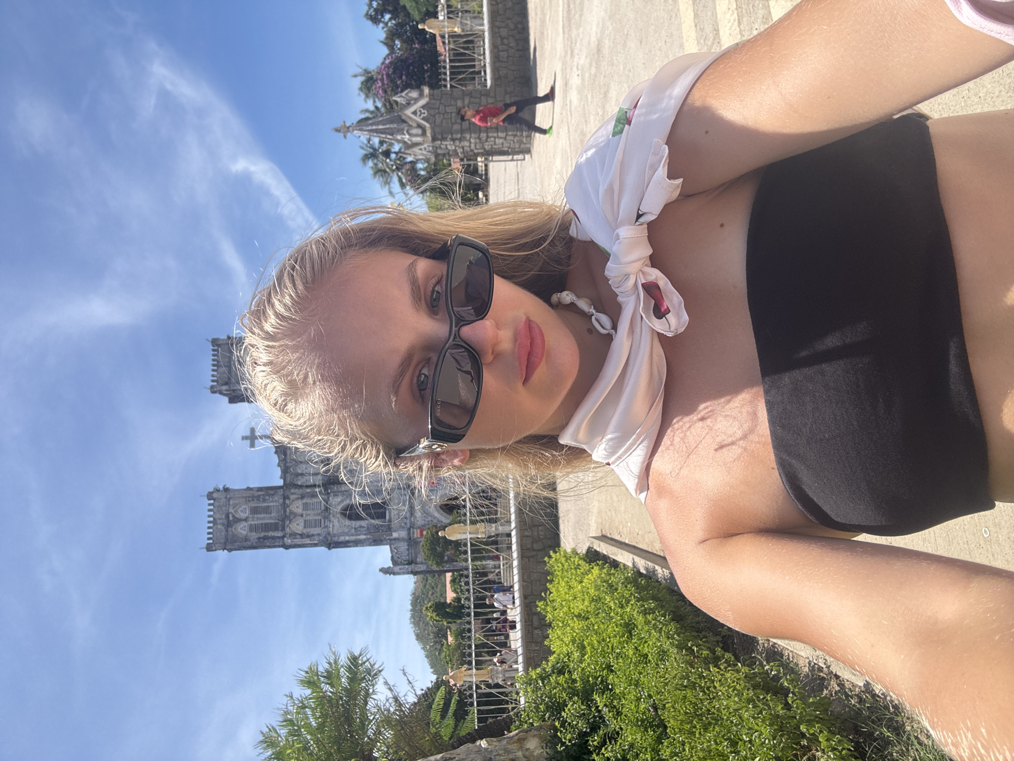 A blonde woman wearing sunglasses, a black tube top, and a white scarf tied around her shoulders takes a selfie outdoors. She is in front of a large, old stone church with two towers and a cross on top. The church appears to be made of grey stone and has Gothic architectural elements. Behind the church, there are palm trees and a clear blue sky with some wispy white clouds. To the left of the church, there is a well-maintained green hedge and a stone pathway. In the background, several people are walking around the church grounds, and one person in a red shirt is walking away from the camera near an entrance to the church. The overall setting suggests a sunny day in a historical or religious site. This location is identified as the Basilica of the Immaculate Conception in Hội Tín, Vietnam.