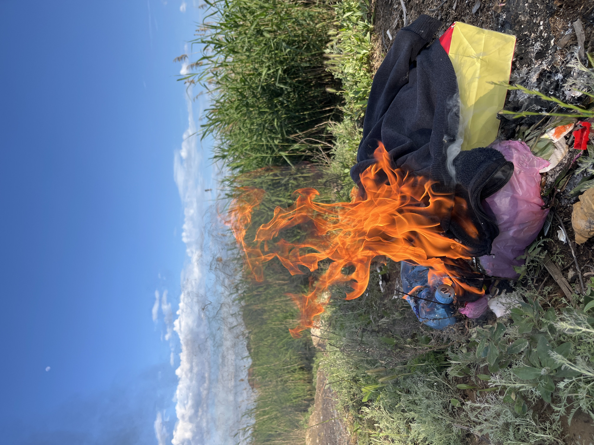 A fire consumes assorted trash, including what appears to be a dark-colored sweatshirt, plastic bags (pink and blue), and a yellow box. The fire is set amongst dry grass, low-lying green plants, and taller grass reeds. Ash and partially burned debris surround the fire. A blue sky with white clouds is visible in the background. The location is identified as Kaspiysk, Russia.