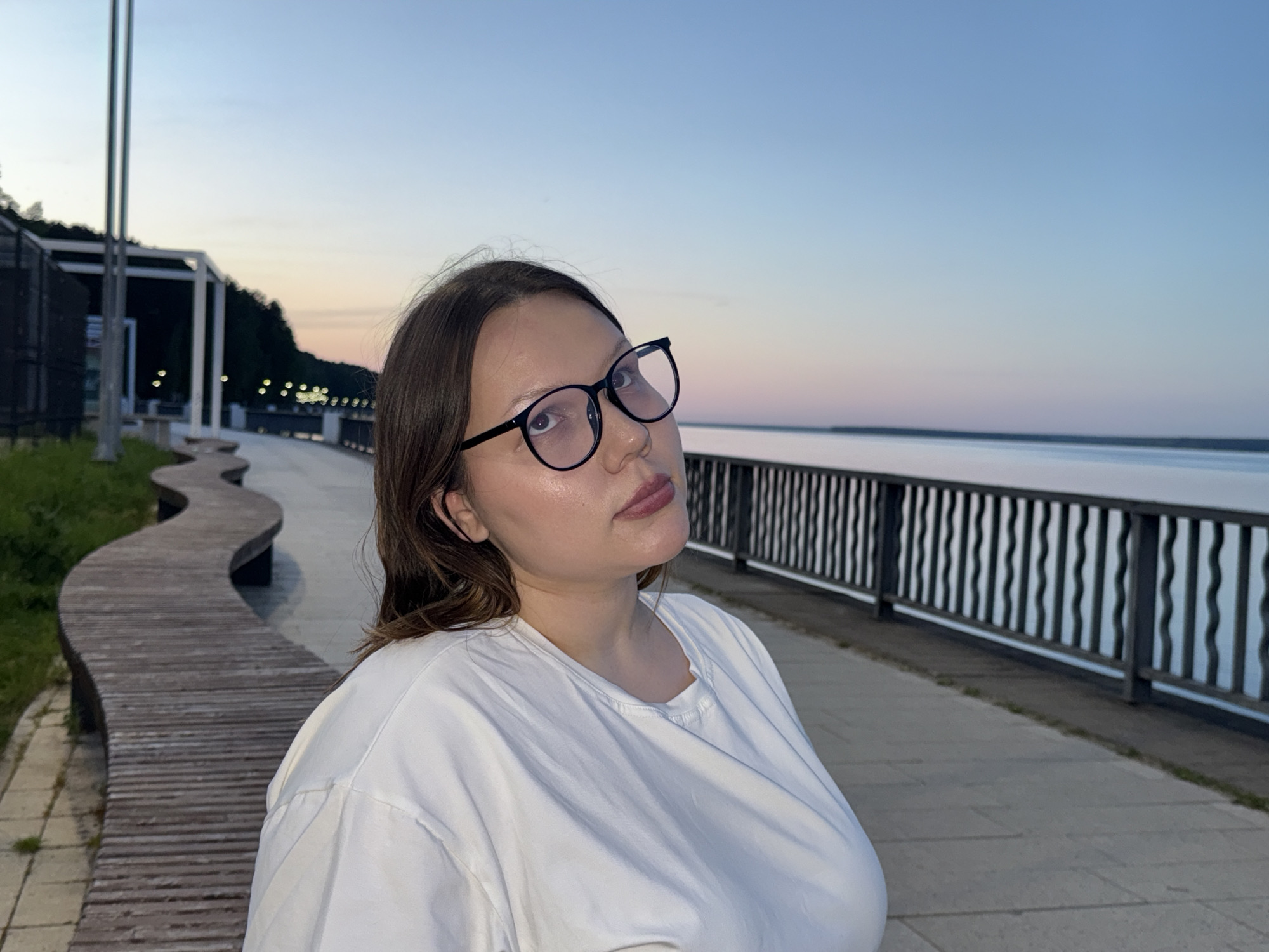 The photograph shows a young woman with light brown hair and glasses standing outdoors, likely in a public space. She is wearing a white t-shirt. The background suggests an urban waterfront setting at dusk or dawn. A winding wooden bench is visible to the left, and a metal railing separates a paved walkway from the water on the right. In the distance, the water stretches towards a horizon with a faint gradient of color in the sky, transitioning from blue to a subtle pinkish hue. The scene is in Chaikovsky, Russia, based on the bench design characteristic of the town's public spaces.