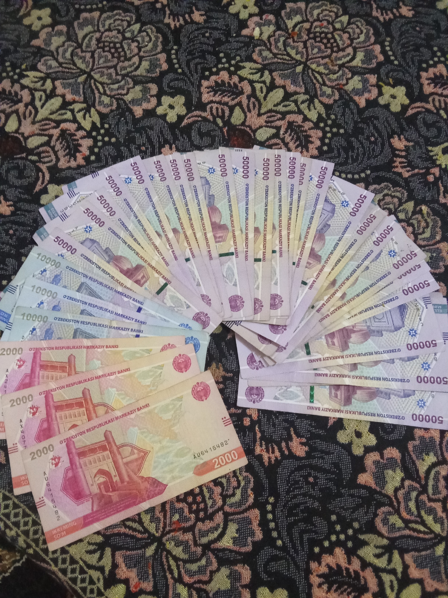 The image shows a fan of Uzbekistani Som banknotes spread out on a patterned fabric. The denominations visible are 50,000 Som, 10,000 Som, and 2,000 Som. The fabric has a dark background with a floral pattern in shades of green, yellow, and pink. The banknotes are arranged in a semi-circular fanned pattern, with the 50,000 Som notes on top, followed by 10,000 Som, and then 2,000 Som notes at the bottom. The image is a top-down view of the banknotes and the fabric. There are no people visible in the image. The specific location in Fergana City, Uzbekistan, is not identifiable from the visual information provided.