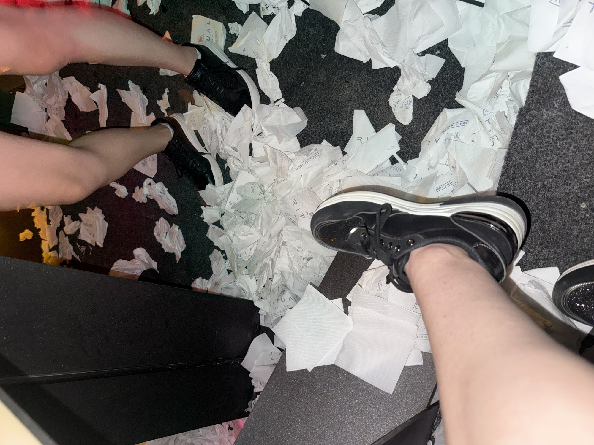 The image shows a floor covered in crumpled white paper. Two individuals are visible, their legs and feet wearing black sneakers. The person in the foreground is wearing one black sneaker and a portion of their leg is visible. The person in the background has both legs and feet visible, also wearing black sneakers. The environment appears to be indoors, possibly a club or event space, given the debris on the floor. No specific details about the city of Sofia, Bulgaria are present in the image.
