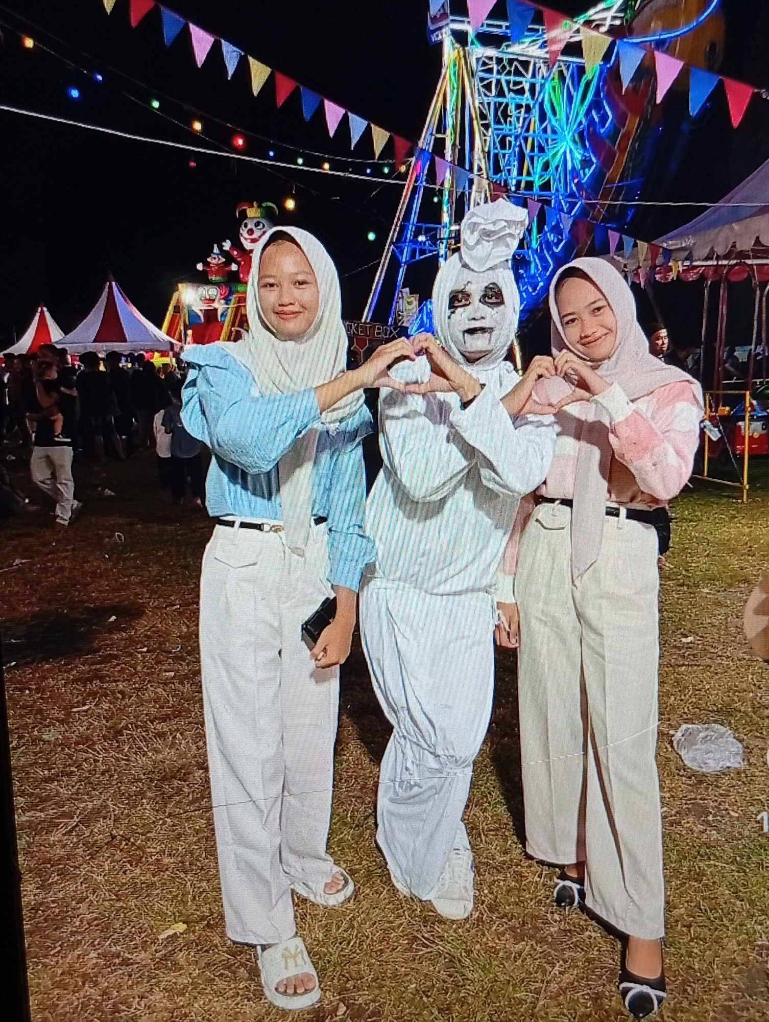 The image depicts three individuals posing for a photograph at a nighttime fairground in Lhokseumawe, Indonesia. The sky is dark, illuminated by various artificial lights from rides and string decorations. In the foreground, from left to right: 1. A female individual wears a light-colored hijab, a light blue long-sleeved top with subtle vertical stripes or texture, and white long pants. She has a black belt with a gold-colored buckle and wears white slider sandals with a dark "NY" logo. Her right hand holds a black smartphone, and both hands are positioned to form the left half of a heart shape. She is smiling and looking towards the camera. 2. A person in the center wears a full white costume, resembling a ghost or mummy. The head is covered with white fabric, possibly bunched like a chef's hat, and the face is painted white with dark, heavy makeup around the eyes and black lines extending from the mouth, creating a skeletal or ghoulish appearance. This individual's hands are positioned to form the central part of the heart shape, completing it with the adjacent subjects' hands. White sneakers are visible on their feet. 3. A female individual wears a light pink or peach-colored hijab, a long-sleeved top featuring patches or stripes of pink and white, and light beige or cream-colored long pants with a subtle vertical seam or slit on the front of the left leg. She has a thin black belt and wears black flat shoes with white bow-like details. Her hands are positioned to form the right half of the heart shape. She is smiling and looking towards the camera. The collective hand gesture of the three individuals forms a complete heart shape. In the midground and background, various fair attractions are visible. Overhead, colorful triangular bunting (pennant flags) in red, pink, yellow, blue, and green, along with string lights, crisscross the dark sky. To the far left, multiple red and white striped conical tents are present, with a crowd of people moving between them. Slightly to the right of these tents, a large, colorful inflatable or statue of a clown character with a red nose and green crown stands on a platform. To the far right, a large Ferris wheel dominates the scene, brightly lit with blue, green, and yellow neon-like lights. Parts of a red and white tent structure and a partially visible "TICKET BOX" sign are also