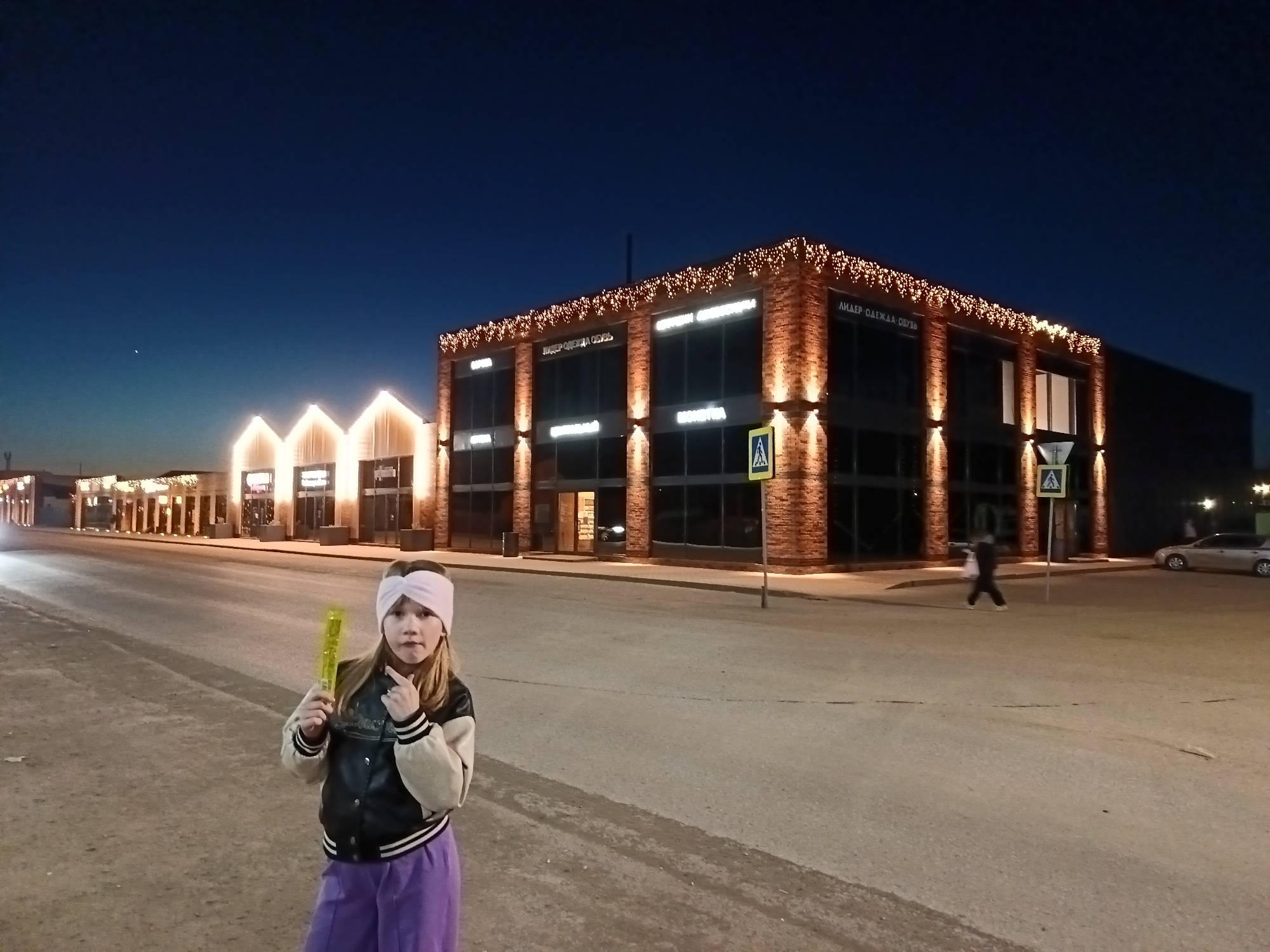 At night in Osa, Russia, a young girl wearing a pink headband, black and white jacket, and purple pants stands on an empty street. She holds a yellow, glowing stick and points to her head with her index finger. Behind her are several illuminated buildings with lighted Christmas decorations along their rooftops. A pedestrian crosses the street behind her. A car is parked on the right side of the street. Streetlights and signs are visible. The sky is dark blue.