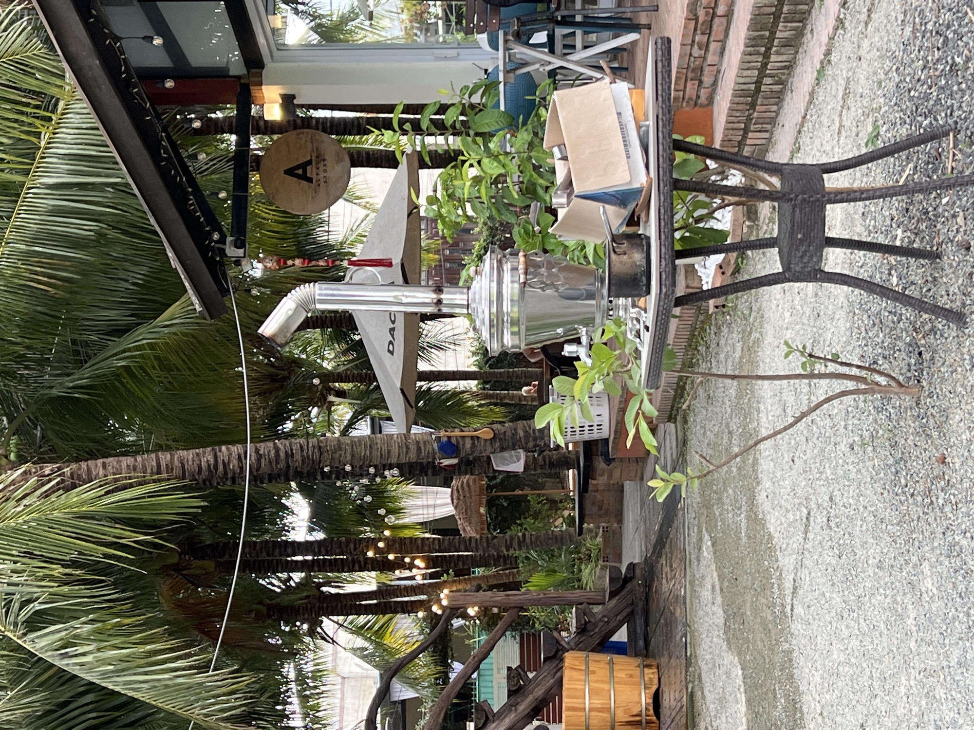 A cafe scene in Tây Nha Trang, Vietnam. A table with a samovar is surrounded by lush vegetation and palm trees. Overhead string lights. A small sign with the letter "A" in the background. Crushed stone on the ground to the right, and a brick border alongside the table.