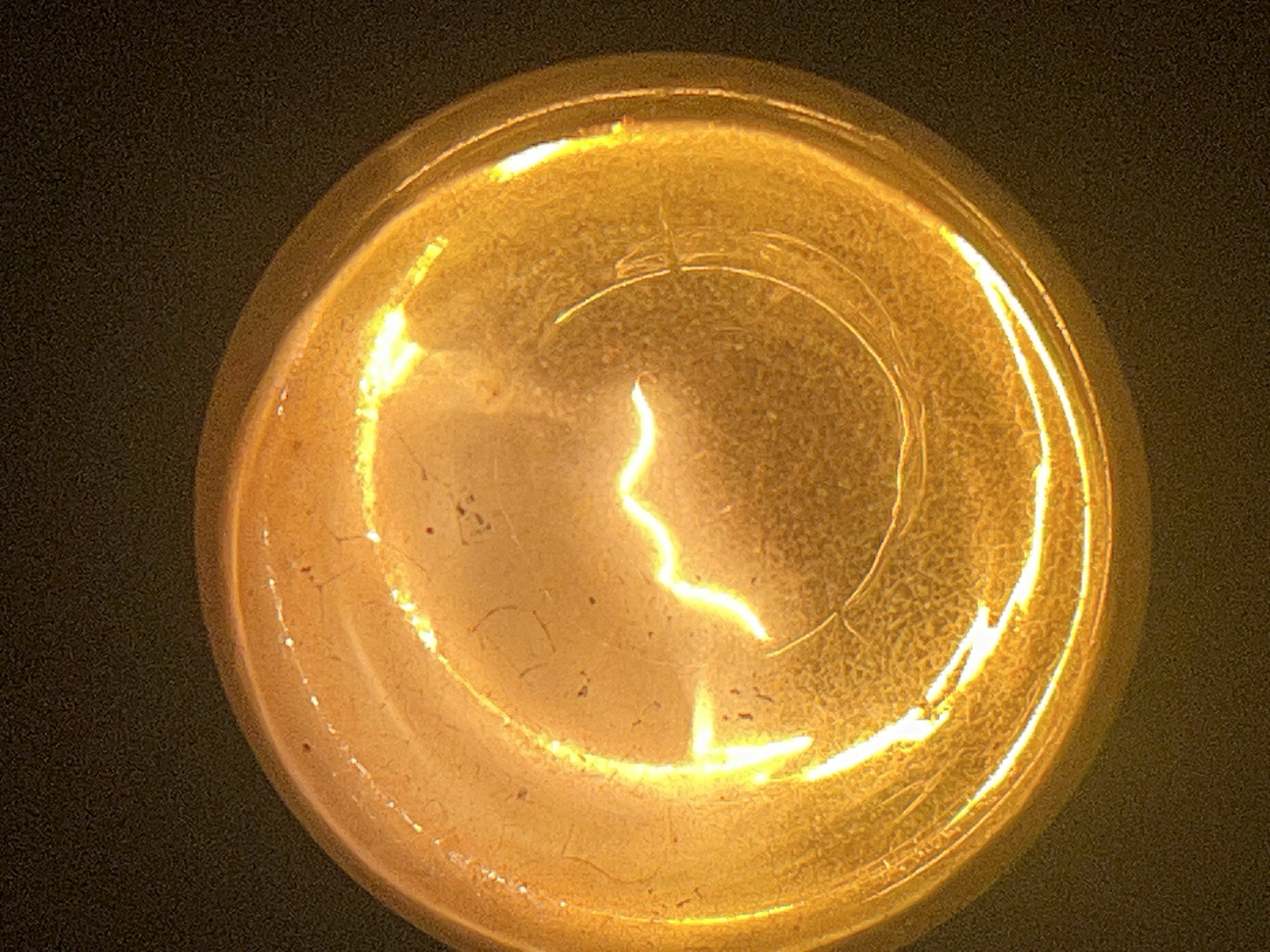 A close-up, overhead shot of a lit incandescent light bulb against a dark background. The bulb is oriented with its base towards the bottom of the frame and its glass enclosure facing upwards, with the filament clearly visible and glowing brightly. The filament is a thin, wavy wire, emitting a warm, yellow-orange light. The glass of the bulb appears to have a textured or frosted surface, with subtle imperfections and reflections visible. The overall lighting is dim, with the light bulb being the primary light source, creating a strong glow that illuminates the surrounding glass and casts a soft light on the dark background. The image captures the intimate detail of a functional object, highlighting its internal structure and the light it produces.