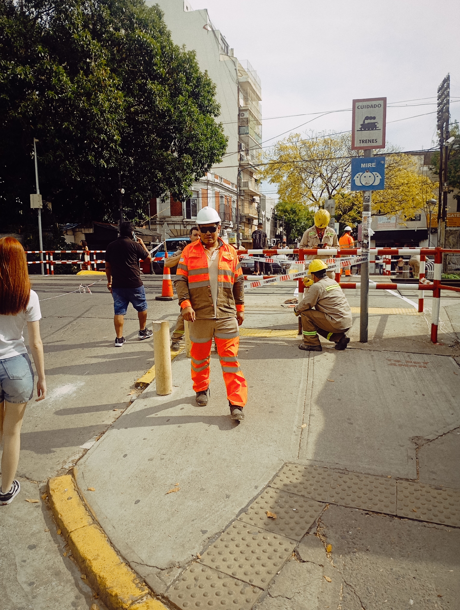 The image shows a street scene in Buenos Aires, Argentina, with a group of construction workers near a railroad crossing. A woman with red hair is walking away from the camera on the left side of the frame. A man in an orange and grey reflective jumpsuit and a white hard hat is standing prominently in the foreground, facing the camera. Behind him, another man is walking away from the camera in a black shirt and blue shorts.
Further back, two workers in yellow hard hats and tan uniforms appear to be inspecting or working on the railroad tracks, which are blocked by red and white barriers. One worker is kneeling near the tracks. The background includes buildings typical of Buenos Aires, and a sign warns of trains. The sky is overcast. The street appears to be under construction or maintenance.