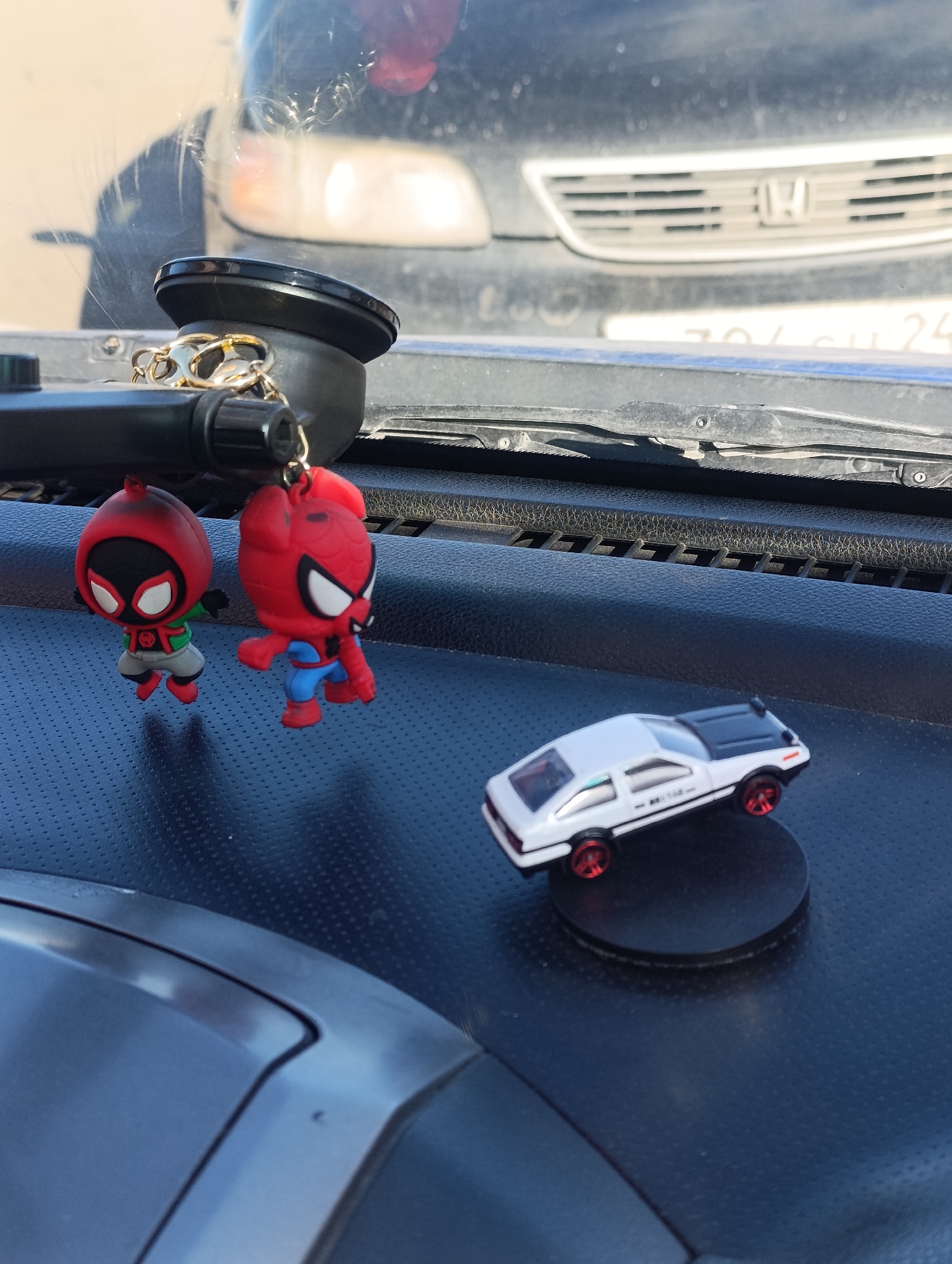The image shows a car dashboard with various items. Hanging from a suction cup mount are two Spider-Man figures. A miniature white and black Toyota AE86 car is mounted on a circular base. The car in the background has a Honda logo on the grill. The license plate reads "704 OU 24". The image was taken in Krasnoyarsk, Russia.