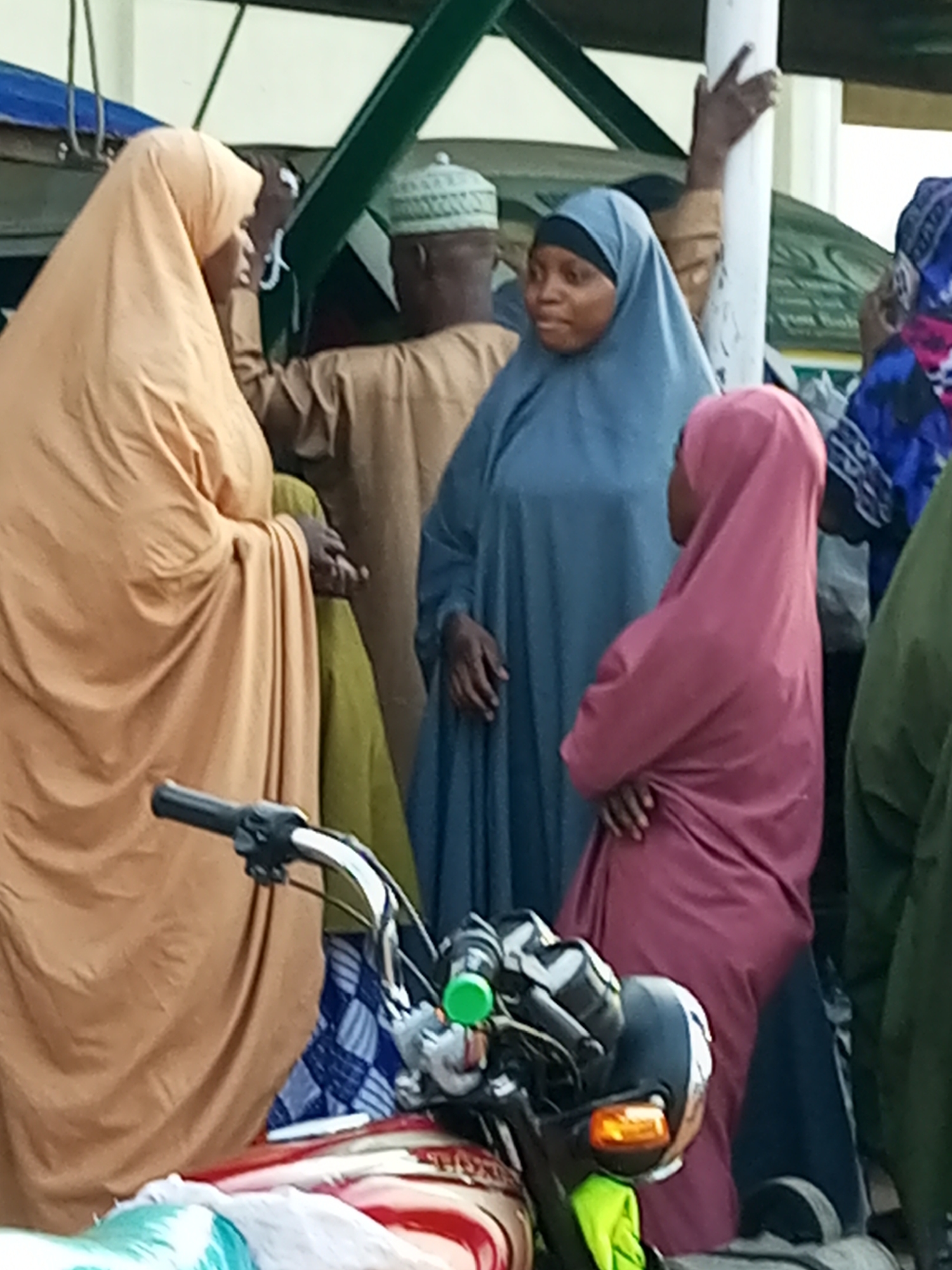 The image depicts a group of individuals, predominantly women wearing hijabs, standing near a white pole and what appears to be a covered structure. In the foreground, the handlebars and part of the front of a motorcycle are visible, suggesting a street scene or public gathering. One woman in a blue hijab is looking to her left, while another in a pink hijab is facing away from the camera. A woman in an orange hijab is on the far left, her head turned down. Behind them, a man wearing a patterned cap and brown clothing is partially visible. The overall impression is a candid street photograph, possibly taken in a busy area of Gombe, Nigeria, given the cultural attire.
