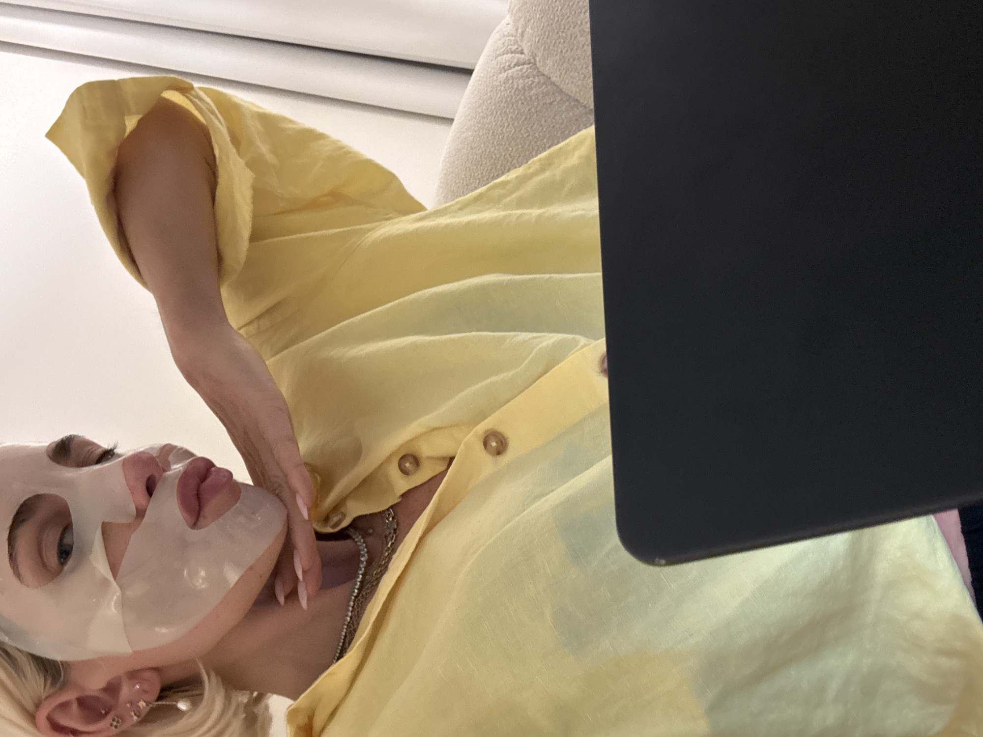 A woman wearing a yellow shirt and a facial mask is looking up at the camera. Her hand is touching her face. She is wearing several necklaces and earrings. A portion of a laptop is visible in the foreground. The scene appears to be indoors. There is no information to confirm this image was taken in Dubai, United Arab Emirates.