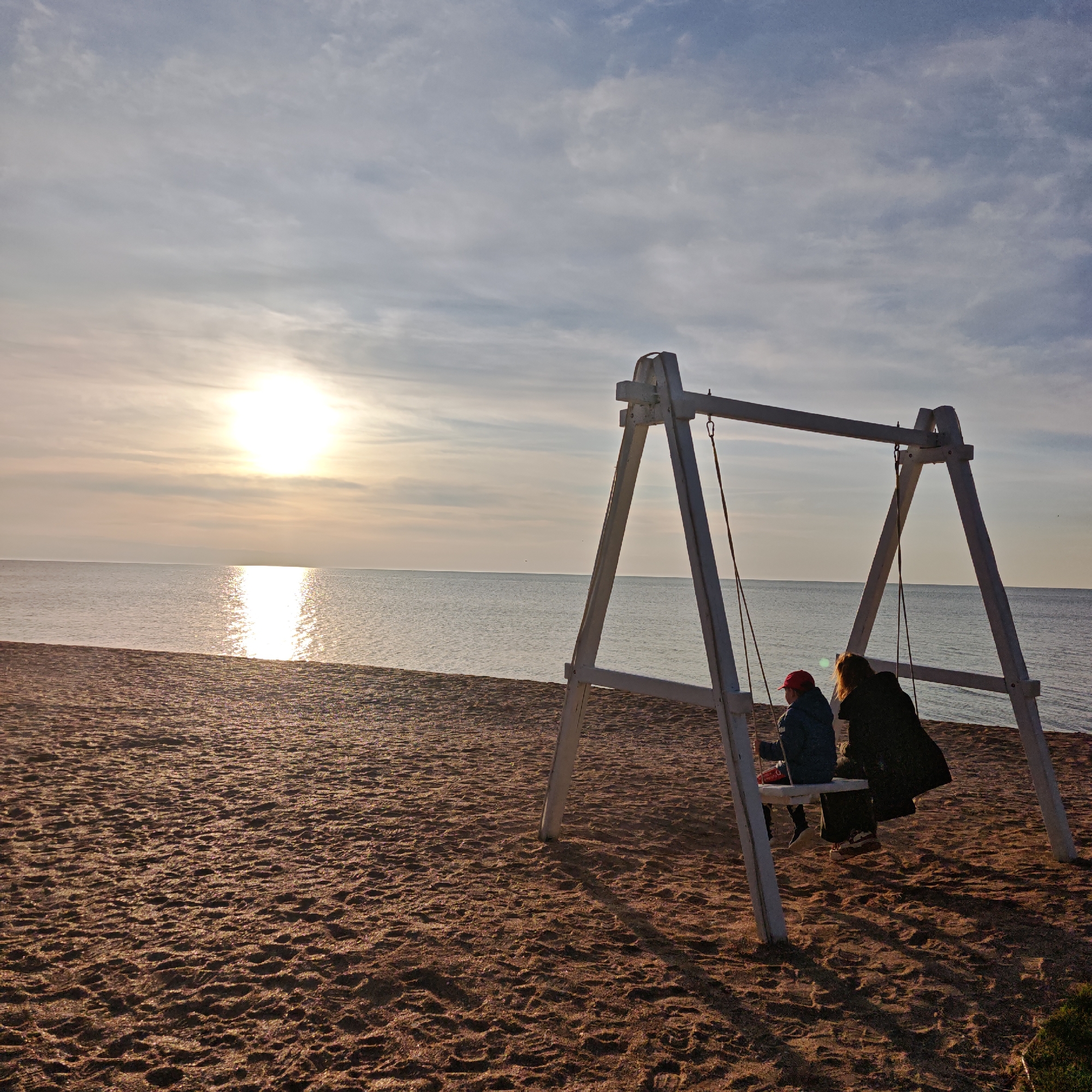 A seaside scene at sunset, location Unknown, Unknown. The sun reflects brightly off the water. A white swing set stands on a sandy beach. Two people are seated on the swing, a child in a red hat and a figure in a dark coat.