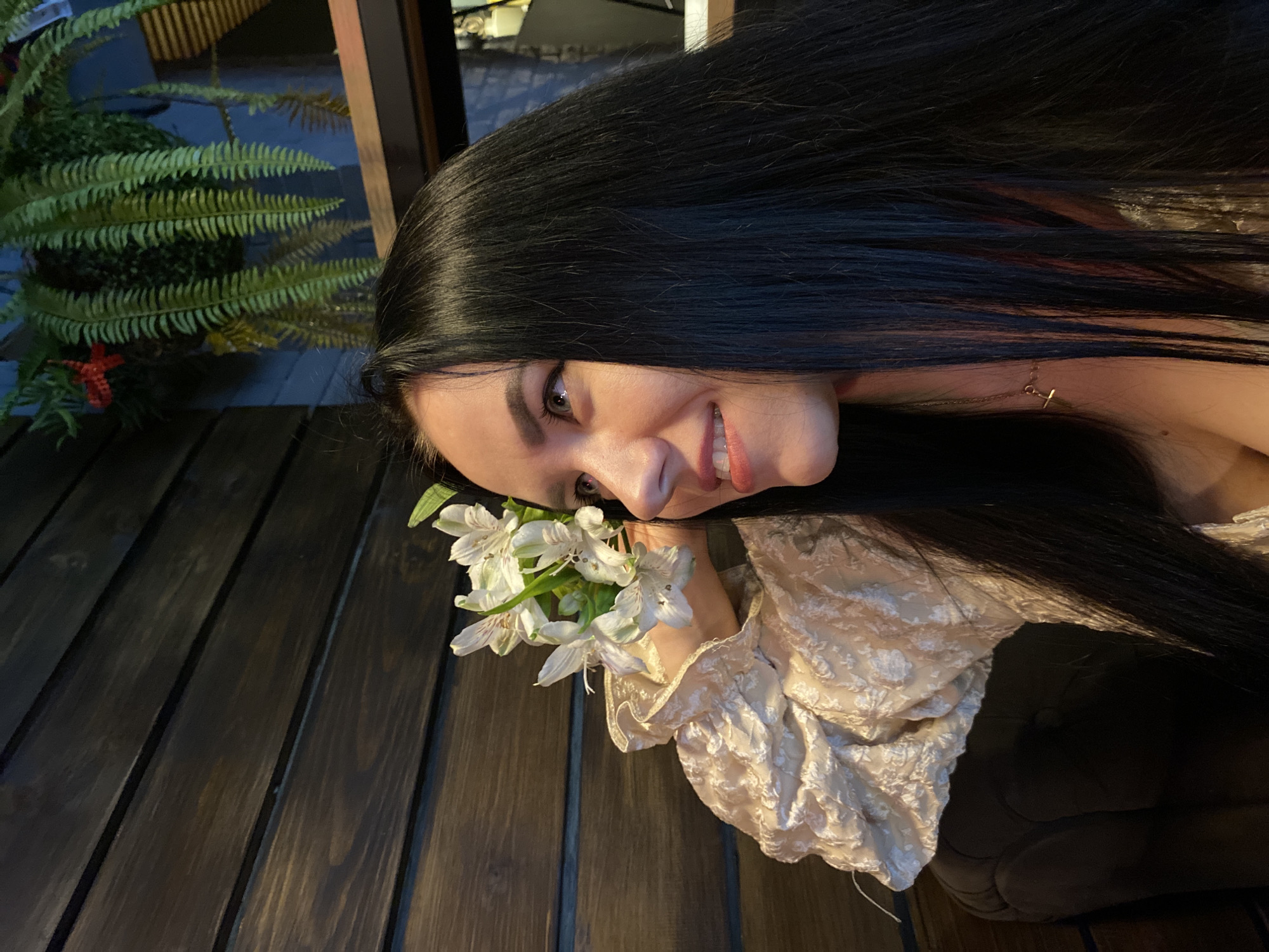 An intimate, rotated portrait captures a young woman with long, lustrous dark hair in a dimly lit setting. She smiles gently, her light-colored eyes (likely hazel or green) looking towards the viewer. Her lips are painted a soft pink, and subtle eye makeup is visible.
She holds a small cluster of white and green flowers, possibly alstroemeria, close to her cheek, partially obscuring her lower face. A delicate golden necklace with a small cross pendant is visible around her neck, peeking through her hair. She wears a light-colored, cream or off-white garment with textured, ruffled fabric, possibly a blouse or dress.
The background features a dark wooden surface with parallel planks, suggesting a table or deck. To the left, a vibrant green fern-like plant is visible, adding natural detail to the scene. The lighting suggests it is evening or night, or an interior space. The overall mood is soft and serene. No text is visible in the image. The location context is Chaikovsky, Russia.