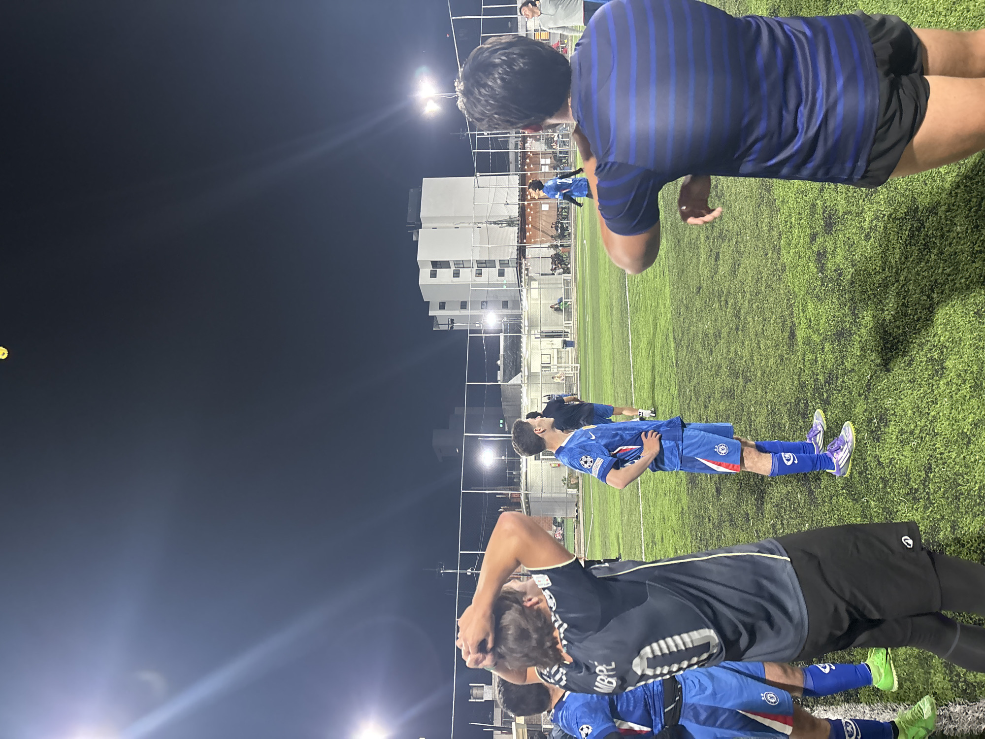 Scene: A group of male soccer players are standing on an artificial turf field at night in San Bernardino Tlaxcalancingo, Mexico. They appear to be stretching or cooling down after a game or practice.
People: The players are wearing soccer jerseys and shorts, with some in blue and others in a navy or black top. Some are wearing compression sleeves or socks.
Environment: A building with lighted windows is visible in the background. The field is illuminated by bright overhead lights. A chain-link fence surrounds the field. The sky is dark.
Objects: Soccer field, artificial turf, building, fence, lights, soccer jerseys, shorts.
Actions: Players are stretching, looking around, or standing with hands on their heads.
Interactions: Players are in close proximity to each other, some appearing to be talking or observing.