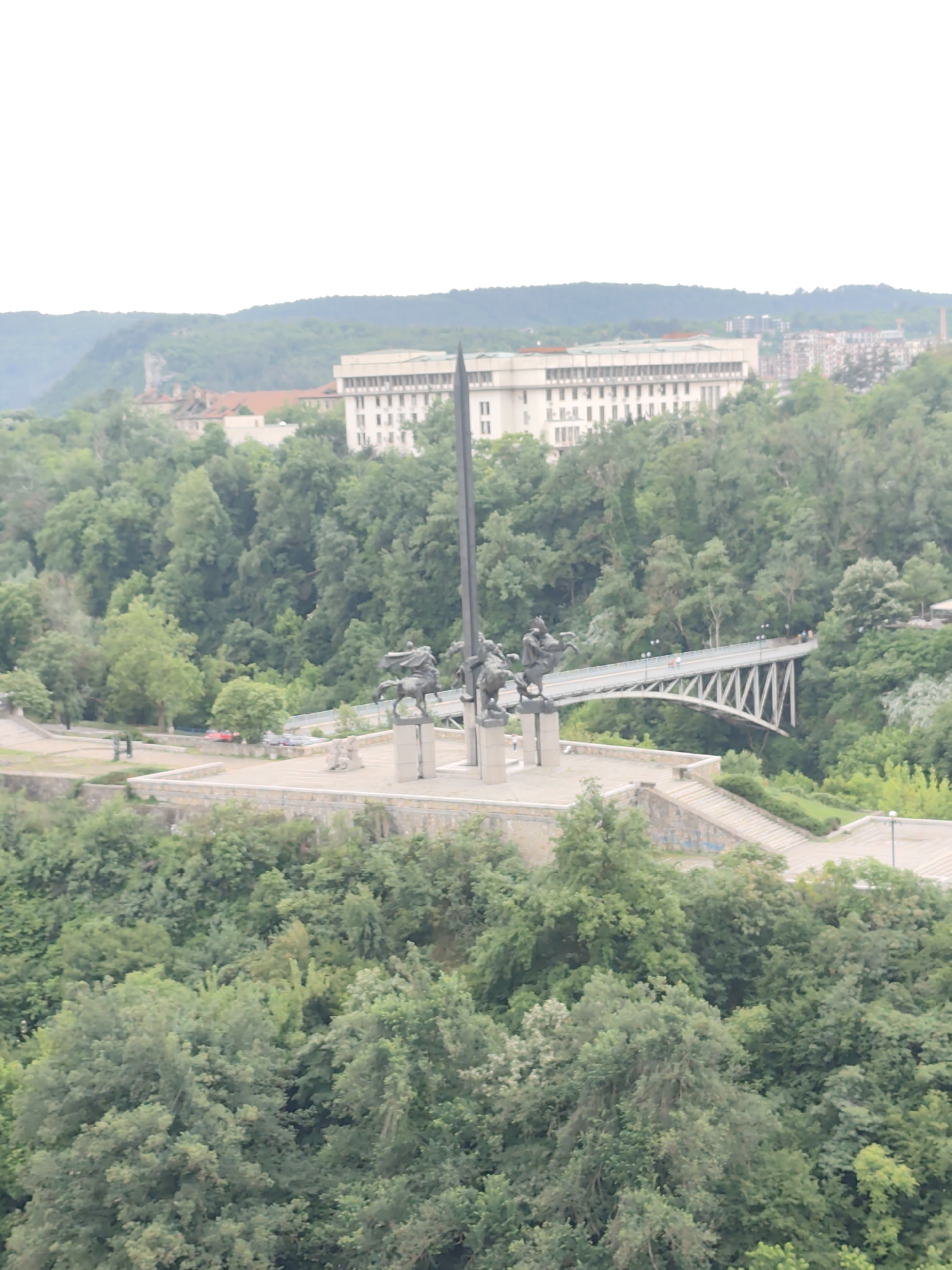 The image shows a distant view of a monument and a bridge located in what appears to be a green, forested area. In the center, a large monument stands prominently, featuring a dark obelisk-like structure with several equestrian statues at its base. A bridge with a distinctive zigzag pattern supports the bridge. The bridge spans a gap between the monument area and the surrounding trees, suggesting it crosses some kind of ravine or valley. The monument and bridge are situated on an elevated platform or clearing, with stairs leading down into the surrounding forest. In the background, a large white building and other structures can be seen on the horizon, indicating the presence of a city or populated area. The environment is dominated by dense trees, which fill the landscape and create a lush, green backdrop. The sky is overcast, giving the scene a muted, soft light. The location is in the city of Unknown, Unknown.