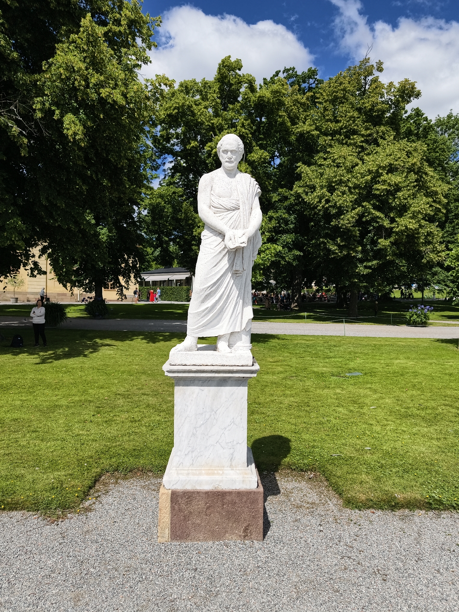 A white marble statue of a man wearing a toga stands on a pedestal in a park. The statue is in the foreground, centered in the frame, casting a shadow to its right. Behind the statue, a lush green lawn stretches out, bordered by a gravel path. In the mid-ground, several people are visible walking and sitting on benches under the shade of large, leafy trees. A building with yellow walls and a dark roof is visible to the left, with more trees and foliage surrounding it. The sky above is blue with scattered white clouds. The statue is located at the Hagaparken park in Ekerö kommun, Sweden.