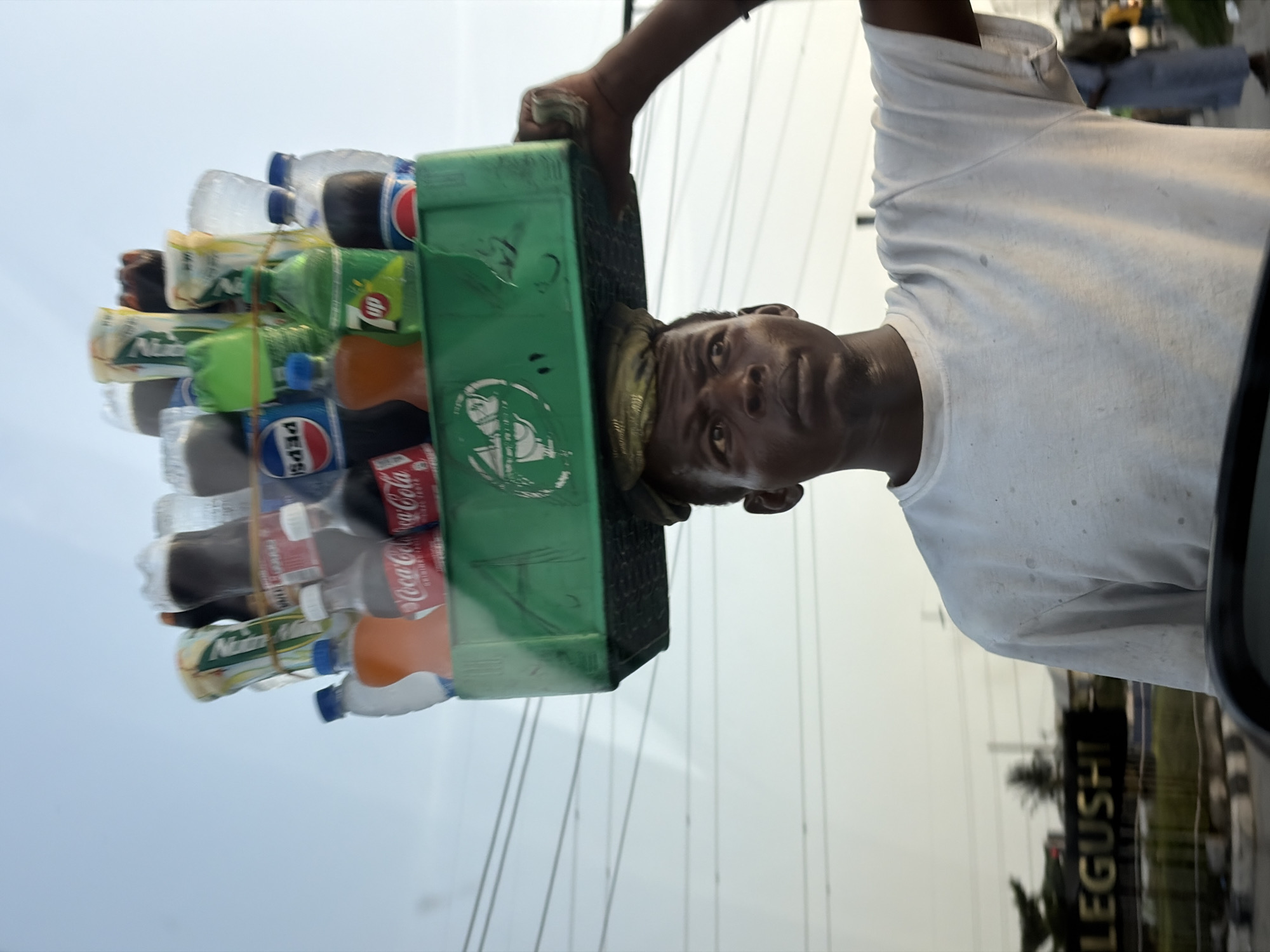 A man in Alaguntan, Nigeria, is balancing a green plastic crate filled with an assortment of bottled drinks on his head. The crate appears to contain Coca-Cola, Pepsi, 7-Up, and Mirinda. He wears a white t-shirt and a fabric head pad to cushion the crate. Power lines stretch across the sky in the background. The scene is viewed from inside a vehicle, with a partial view of the driver's side window and the word "LEGUSHI" visible on a sign outside.