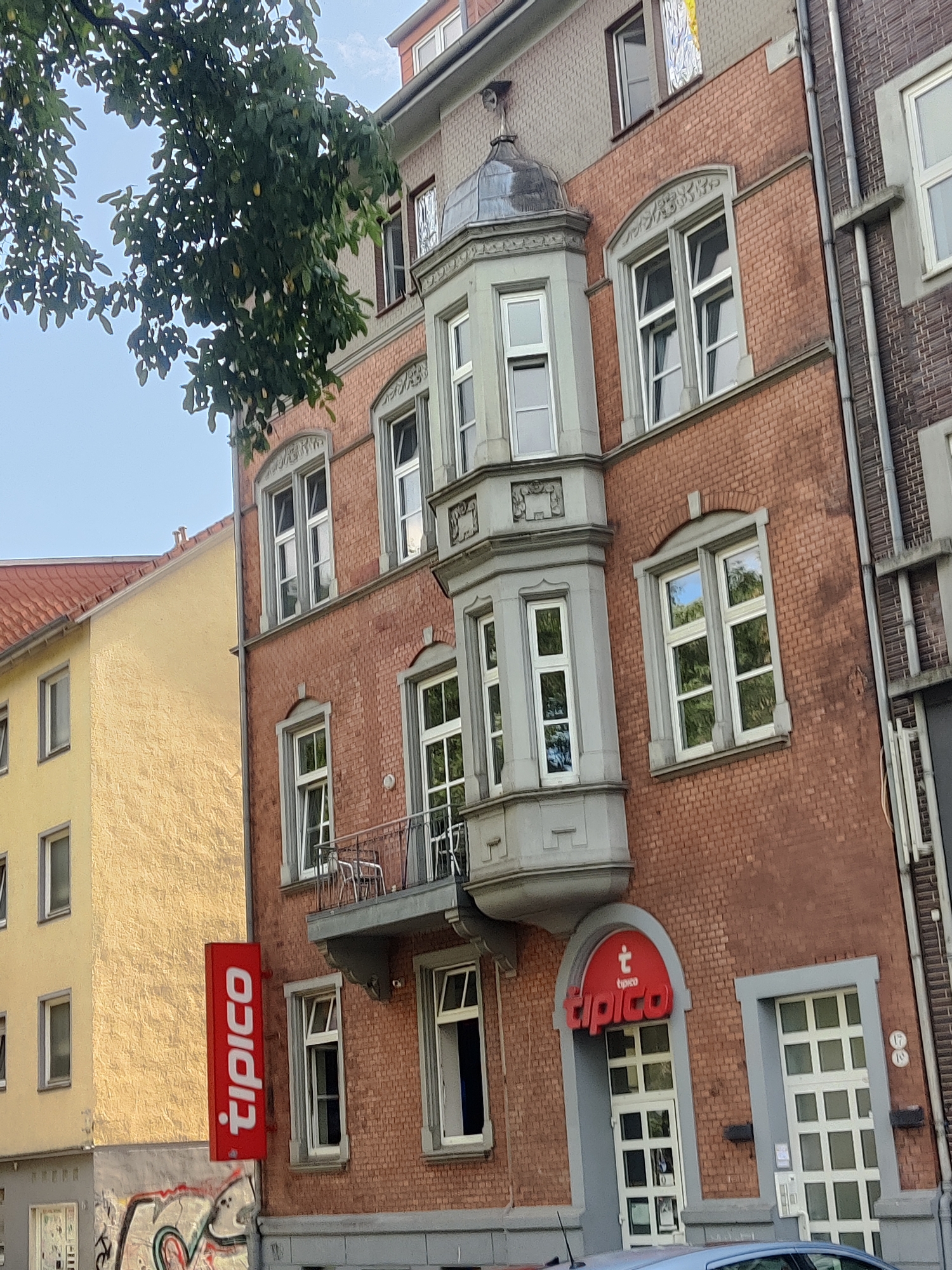 The image shows the exterior of a brick building in Bielefeld, Germany. The building has several windows and a prominent red sign with white lettering that says "tipico". To the left of the brick building is a yellow building with a red roof. The sky is visible in the upper part of the image, and trees with green leaves are in the foreground. The overall scene depicts a street view of urban architecture.