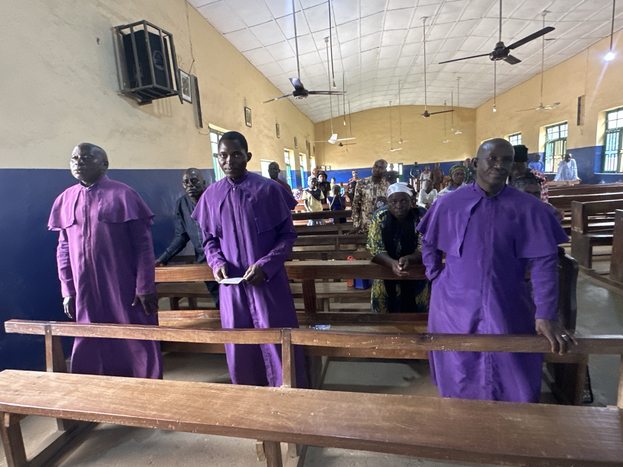 The image depicts the interior of a church in Akwanga, Nigeria. In the foreground, several men are standing, some wearing purple robes. The church has wooden pews and a light-colored ceiling with multiple ceiling fans. The walls are painted in a light yellow color with a blue stripe at the bottom. There are windows on the right side of the room, and a framed picture hangs on the wall. In the background, more people are visible, seated on the pews. The overall impression is of a religious gathering in a modest church setting.