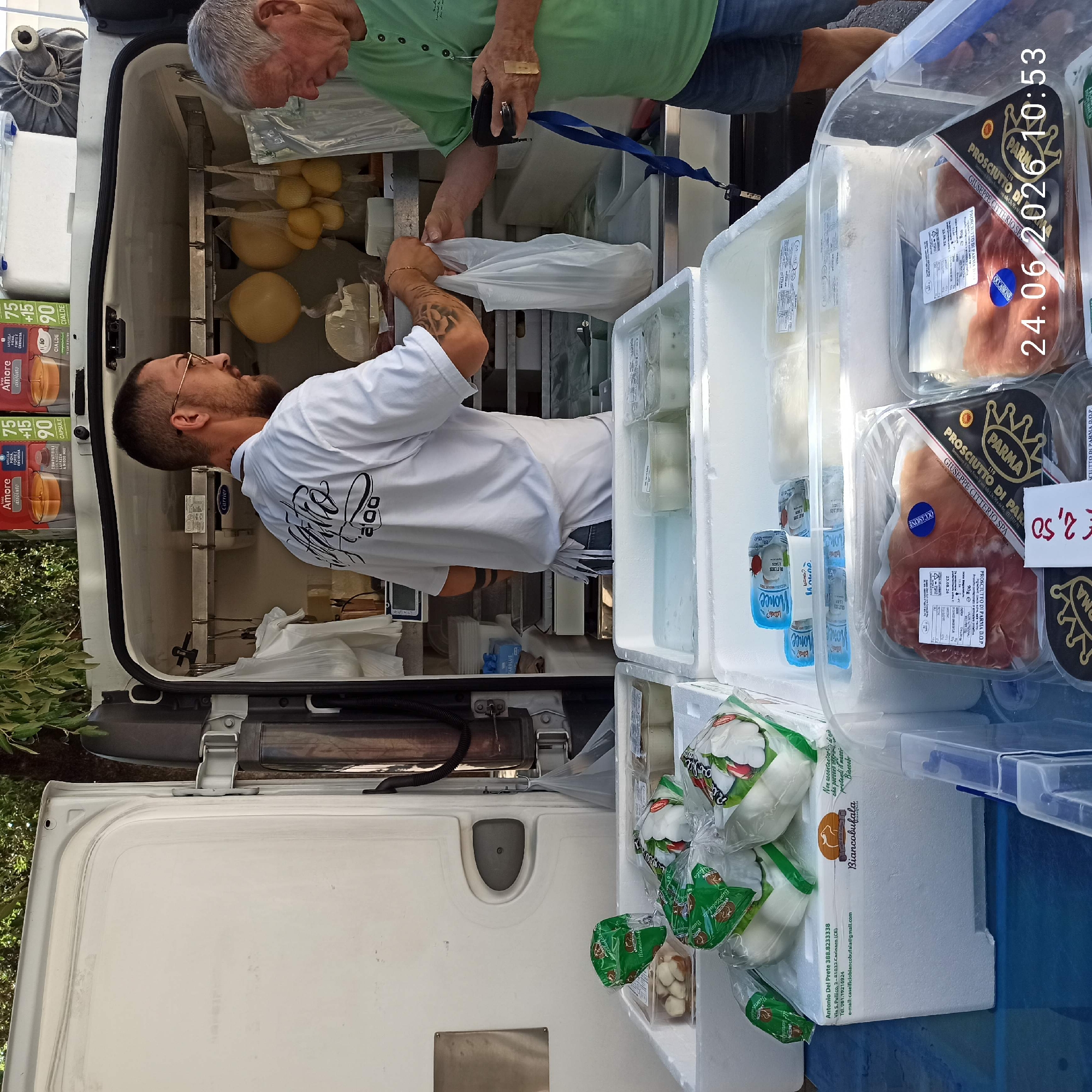 A man in a white t-shirt with glasses is interacting with an older man in a green shirt. The man in the white t-shirt is handing a white plastic bag to the man in the green shirt, who is holding a wallet and a blue lanyard. They are standing in front of a white food truck, which appears to be a mobile cheese stand. Various cheeses are displayed in containers and hanging from hooks inside the truck. Packaged meats, likely prosciutto, are visible in plastic containers in the foreground. The scene is outdoors, with trees and foliage visible in the background. The time stamp "24.06.2026 10:53" is superimposed on the image. The location is identified as Bitonto, Italy.