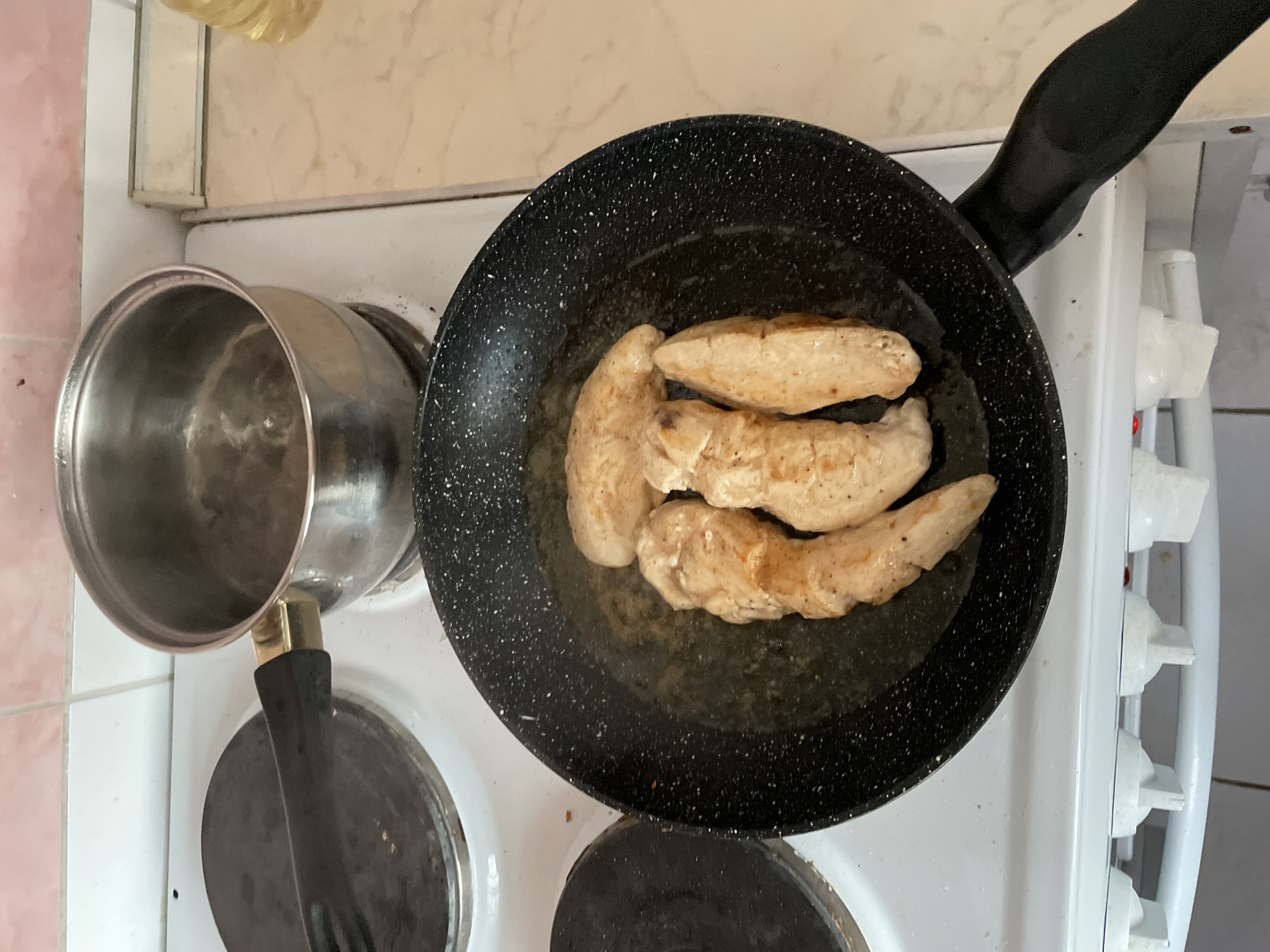 Four pieces of lightly browned chicken tenderloins are being pan-fried in a black, speckled frying pan on a white electric stovetop. A silver pot with water is to the left of the pan. The stovetop is in a kitchen with light-colored countertops and pink tiled backsplash. The scene is consistent with a home kitchen environment in Krasnoyarsk, Russia.