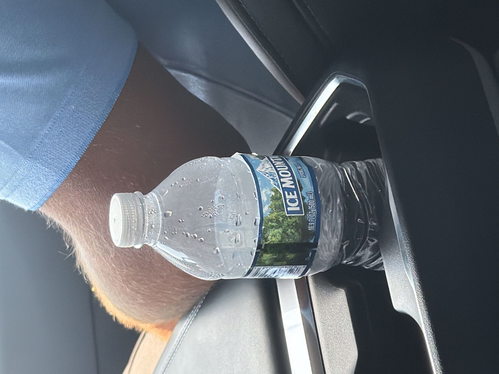 A close-up, eye-level shot captures a partially filled plastic bottle of "ICE MOUNTAIN" spring water resting in a car's center console cup holder. The bottle, condensation beading on its surface, displays a label with an image of a lush green forest and snow-capped mountains. A person's forearm, covered in light brown hair, is visible leaning behind the bottle, partially obscured by the bottle's neck and cap. The person is wearing a light blue polo shirt with the collar visible at the top of the frame. The background consists of the dark interior of a vehicle, including a black leather seat and the textured surface of the center console. The lighting suggests an outdoor environment, likely during the day, with sunlight casting highlights and shadows. The image's context of a car interior suggests it was taken in South Saint Paul, United States of America, although no specific landmarks are visible.
