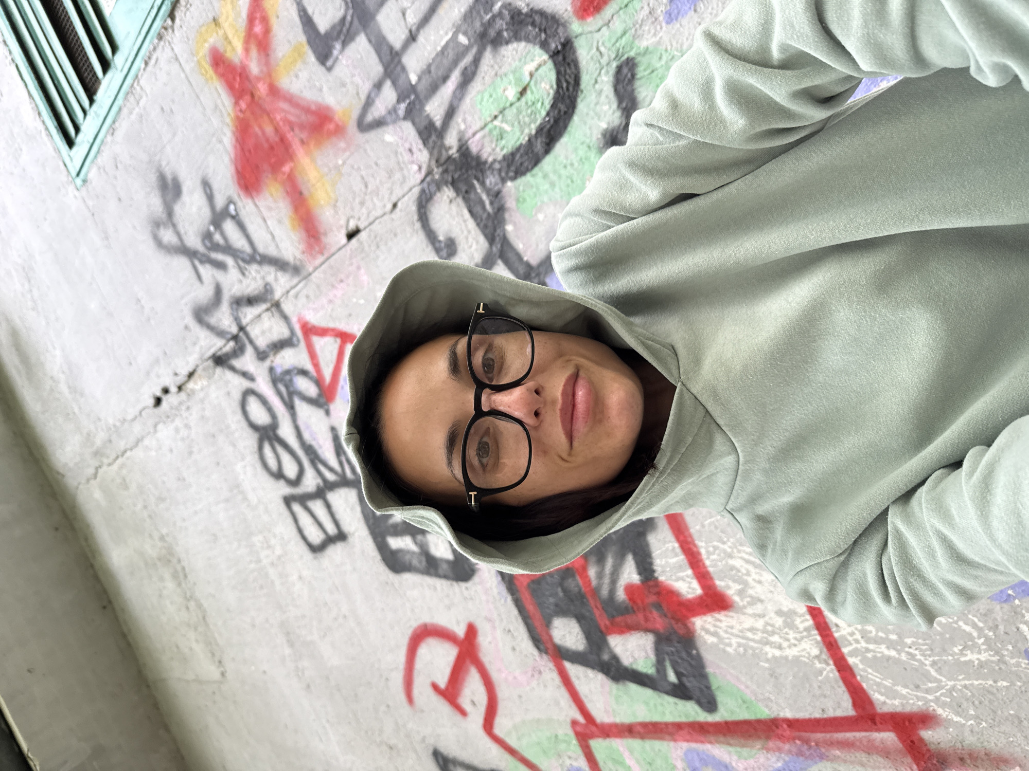A woman with fair skin, wearing black-framed glasses and a light green hooded sweatshirt, is centered in the frame. The background is a concrete wall covered in graffiti, including red, black, and green markings. A blue ventilation window is visible in the upper left corner. The image appears to have been taken in Pleven, Bulgaria, due to the graffiti style being common in the city.