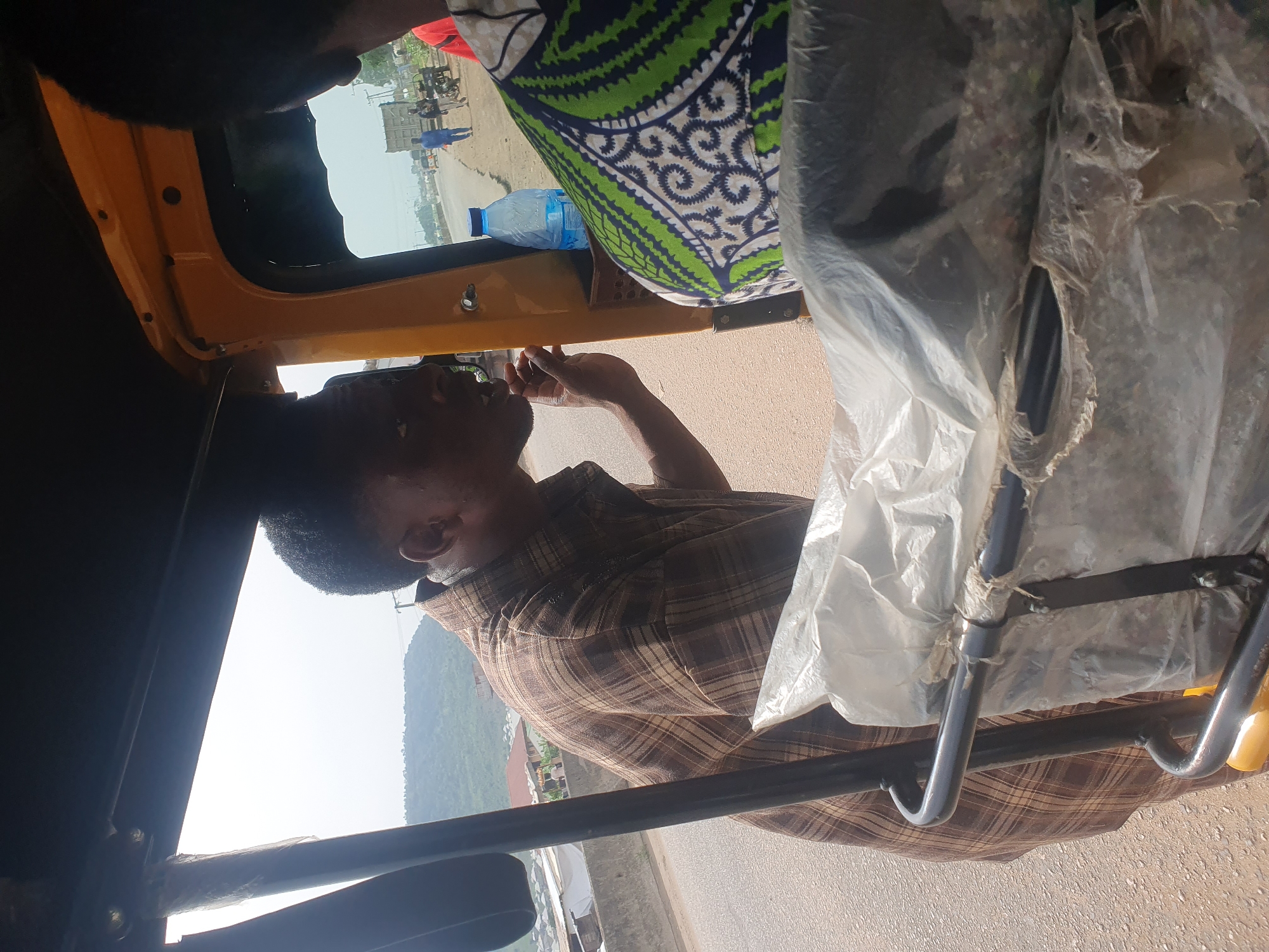 The image depicts the interior of a yellow commercial tricycle, a common form of transportation in Lokoja, Nigeria. The driver, a dark-skinned man with short hair, is visible in the foreground, wearing a plaid shirt. A passenger wearing a green and white patterned fabric sits in the back of the vehicle. A plastic water bottle is located above the driver’s head on the interior shelf of the tricycle. Clear plastic sheeting partially covers the metal frame of the vehicle. Outside the vehicle, a view of a partially clear sky and lush, green hills can be seen.
