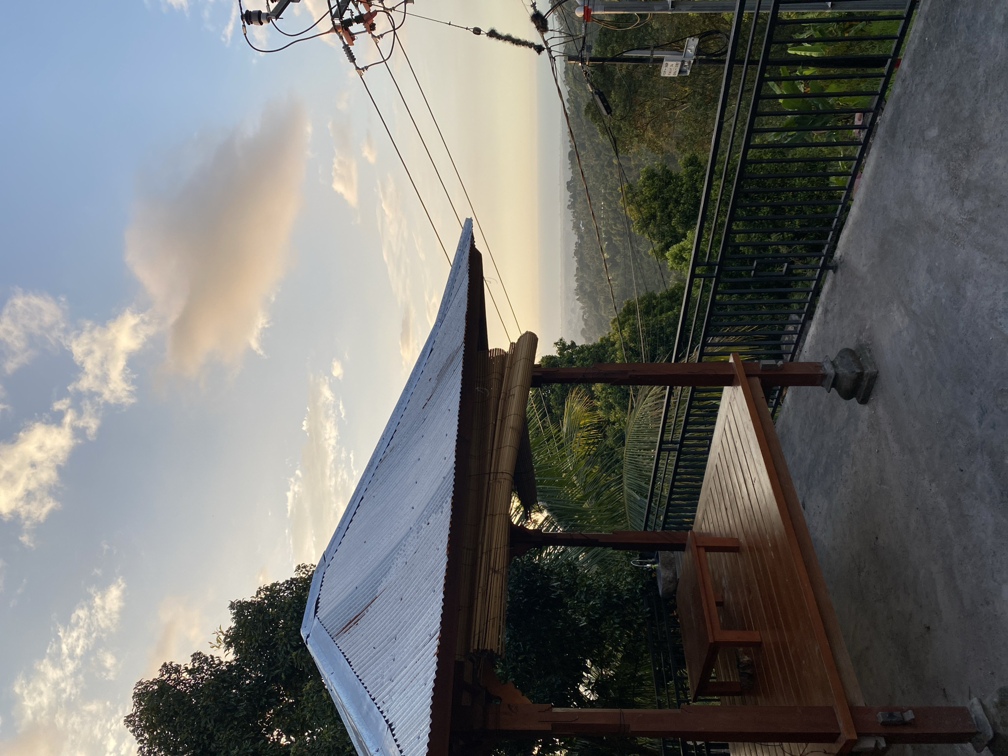 The image depicts a gazeboin Sawan, Indonesia, overlooking a lush, green landscape. The gazeboisconstructed of wood with a corrugated metal roof. Inside, a low wooden table sits on the slatted wooden floor. A black metal railing lines the edge of the elevated platform, beyond which the verdant hills stretch towards the horizon. The sky above is a soft blue, with scattered white and yellow clouds, suggesting either sunrise or sunset. Utility poles and wires are visible on the right side of the frame, indicating the presence of infrastructure in the area. The overall scene is serene and offers a picturesque view of the Indonesian countryside.