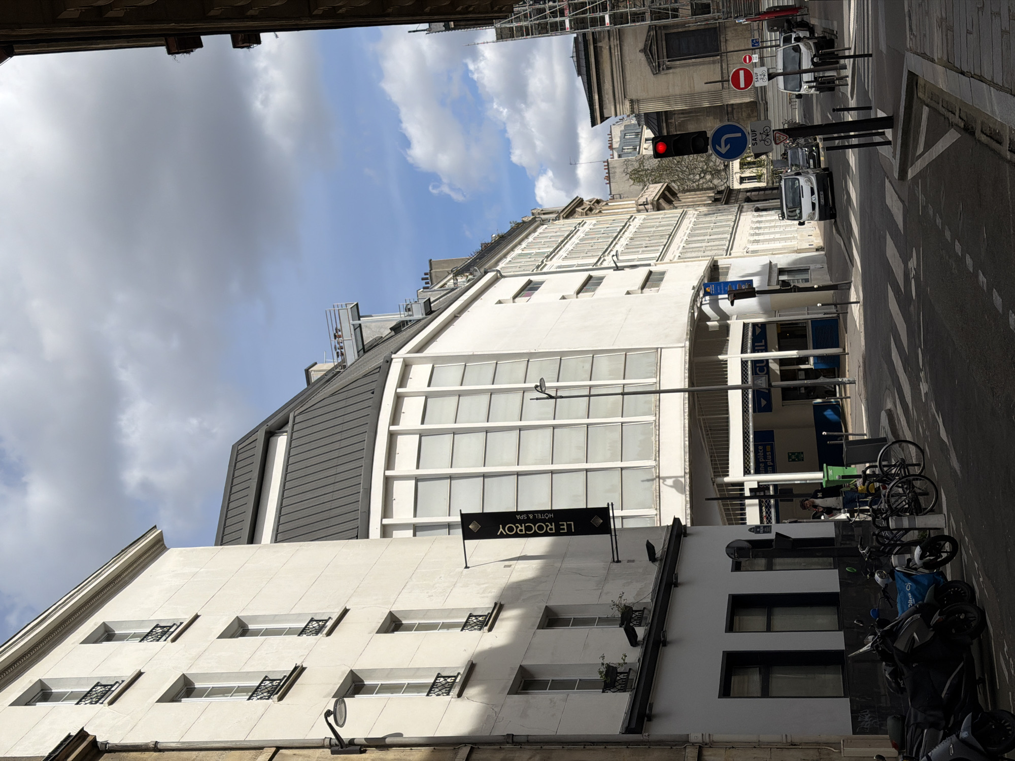 The image depicts a street scene in Paris, France. The focal point is the building displaying the "Le Rocroy Hotel & Spa" sign. The building is predominantly white with a gray roof and features multiple windows. A street runs parallel to the building. On the street, there are parked motorcycles and bicycles, street signs, and a red traffic light. Several people are walking on the sidewalk. The sky is partly cloudy. Buildings are visible across the street. The image is taken from a slightly low angle.
