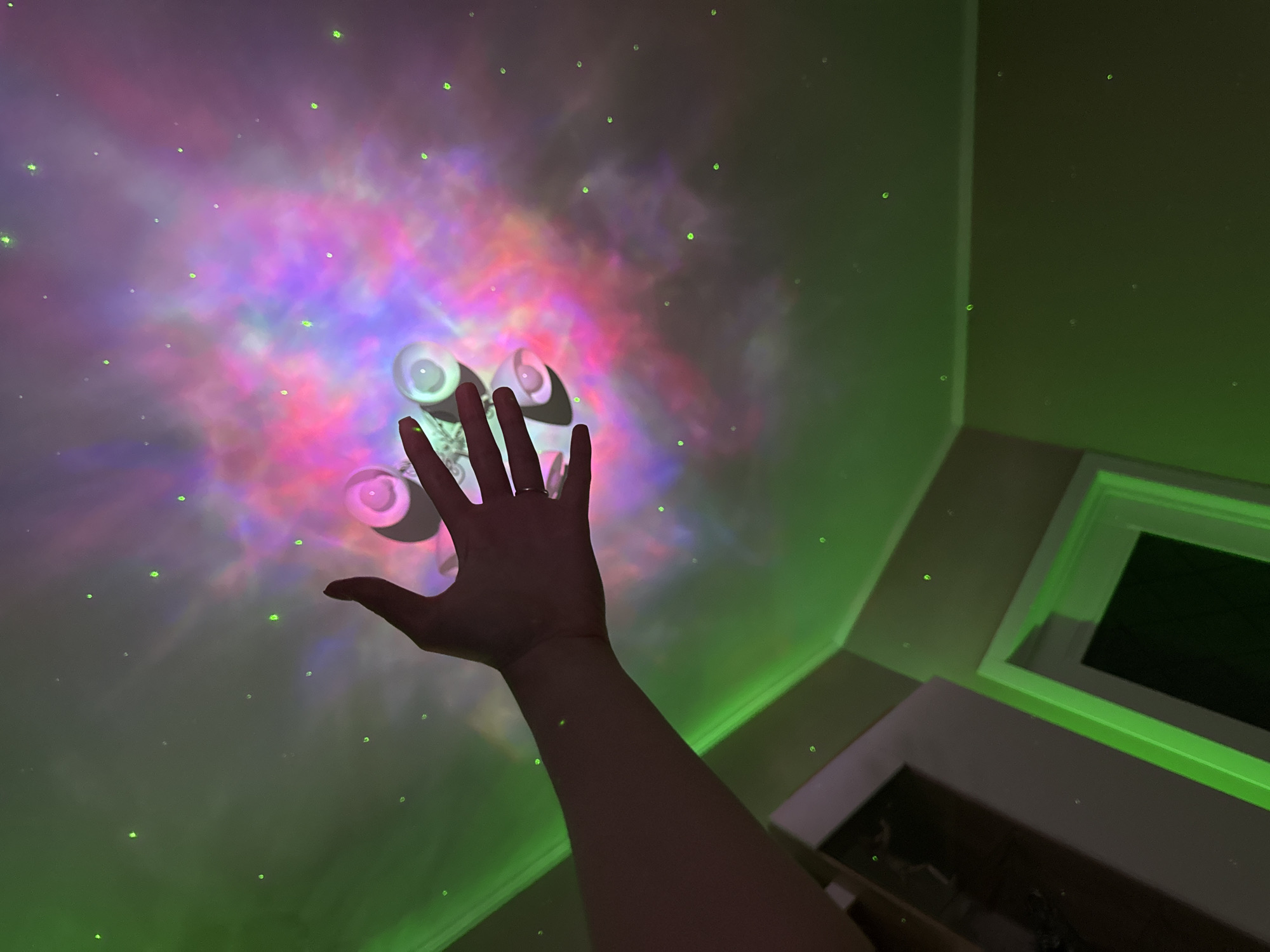 The image depicts a dimly lit room with a ceiling projector casting a nebula-like pattern in shades of pink, purple, and blue, with small green dots resembling stars. A person's left hand is extended towards the projector, which appears to be a small, multi-lensed device attached to the ceiling. Green light also illuminates the corners of the room and parts of the walls. A doorway with a dark interior is visible on the left side of the frame. The overall scene suggests an attempt to create a celestial or cosmic ambiance within a domestic space.
