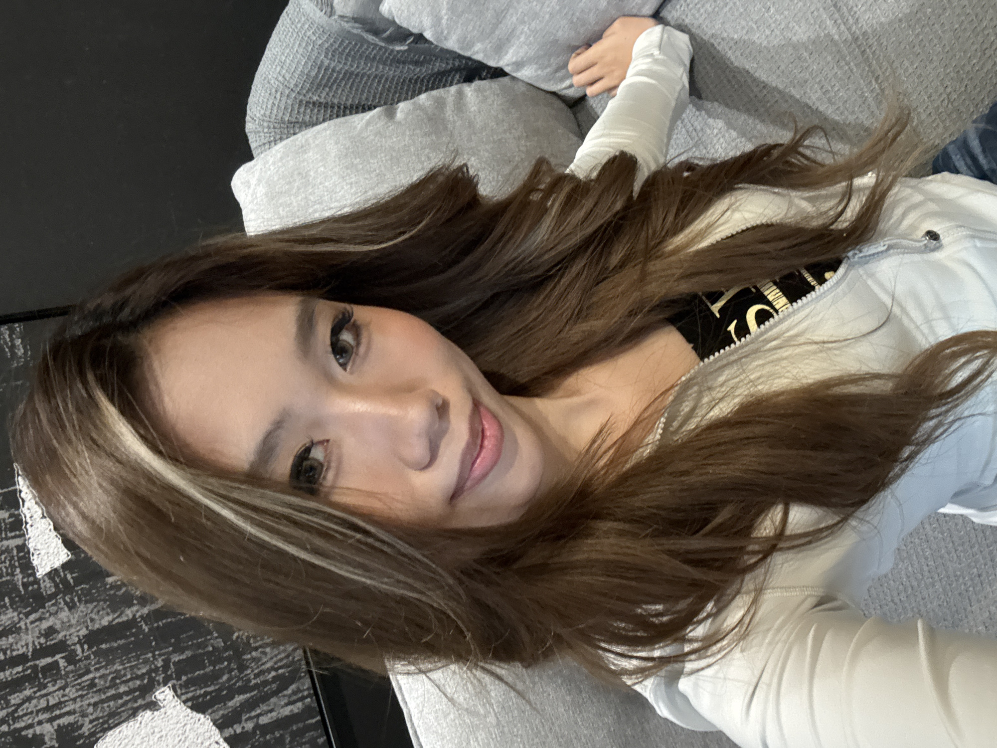 The image is a selfie of an Asian woman with long brown hair, lying on a gray couch. She is wearing a white jacket over a black top with gold lettering. Part of her right arm is covered with a white wrap. The background includes pillows and a dark floor. The location, Liangjiang, China, cannot be verified from the image.