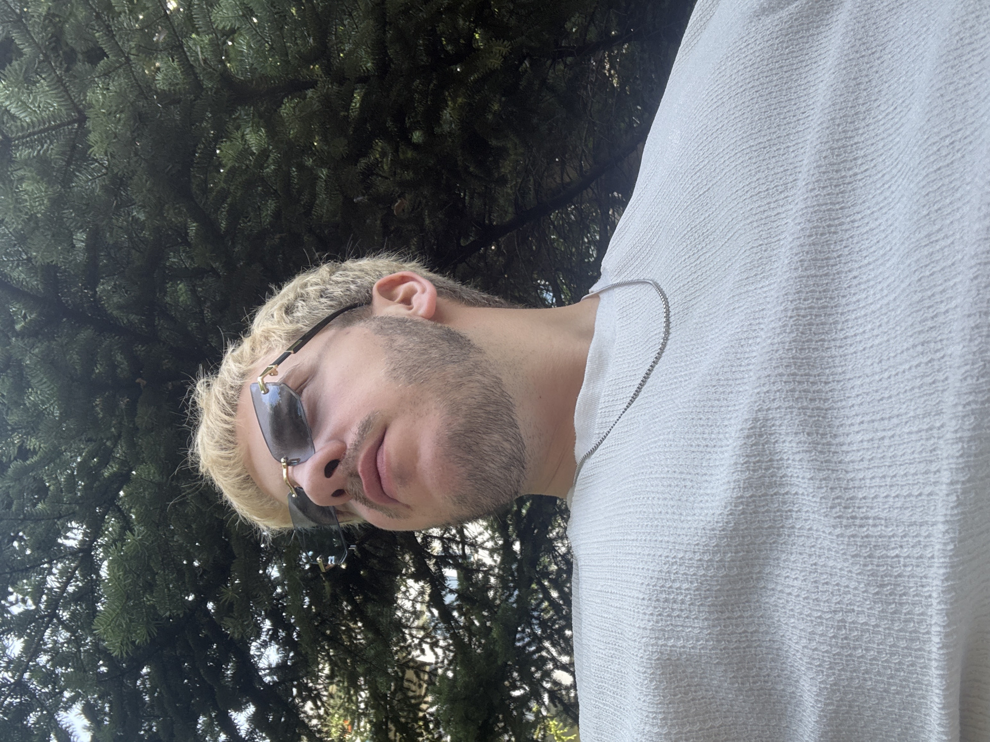The media file presents a portrait-style, low-angle photograph of a person, likely male, from the shoulders up. The person is positioned on the left side of the frame, with their head tilted slightly downward and to the right, looking upwards. They have short, curly blonde hair and a well-groomed stubble or short beard. Dark-tinted sunglasses with gold-colored frames are worn, obscuring their eyes. A thin, silver chain necklace is visible around their neck, partially resting on their light-colored, possibly white, textured shirt. The fabric of the shirt has a distinct, small, rippled or waffle-knit pattern.
The background consists of dense, dark green tree foliage, suggesting an outdoor setting surrounded by evergreen trees like pines or firs. Bright highlights on the leaves indicate sunny weather. The overall impression is a casual, relaxed outdoor moment. There is no discernible activity other than posing for the photograph. The time of day appears to be bright daytime. No text is visible in the image.