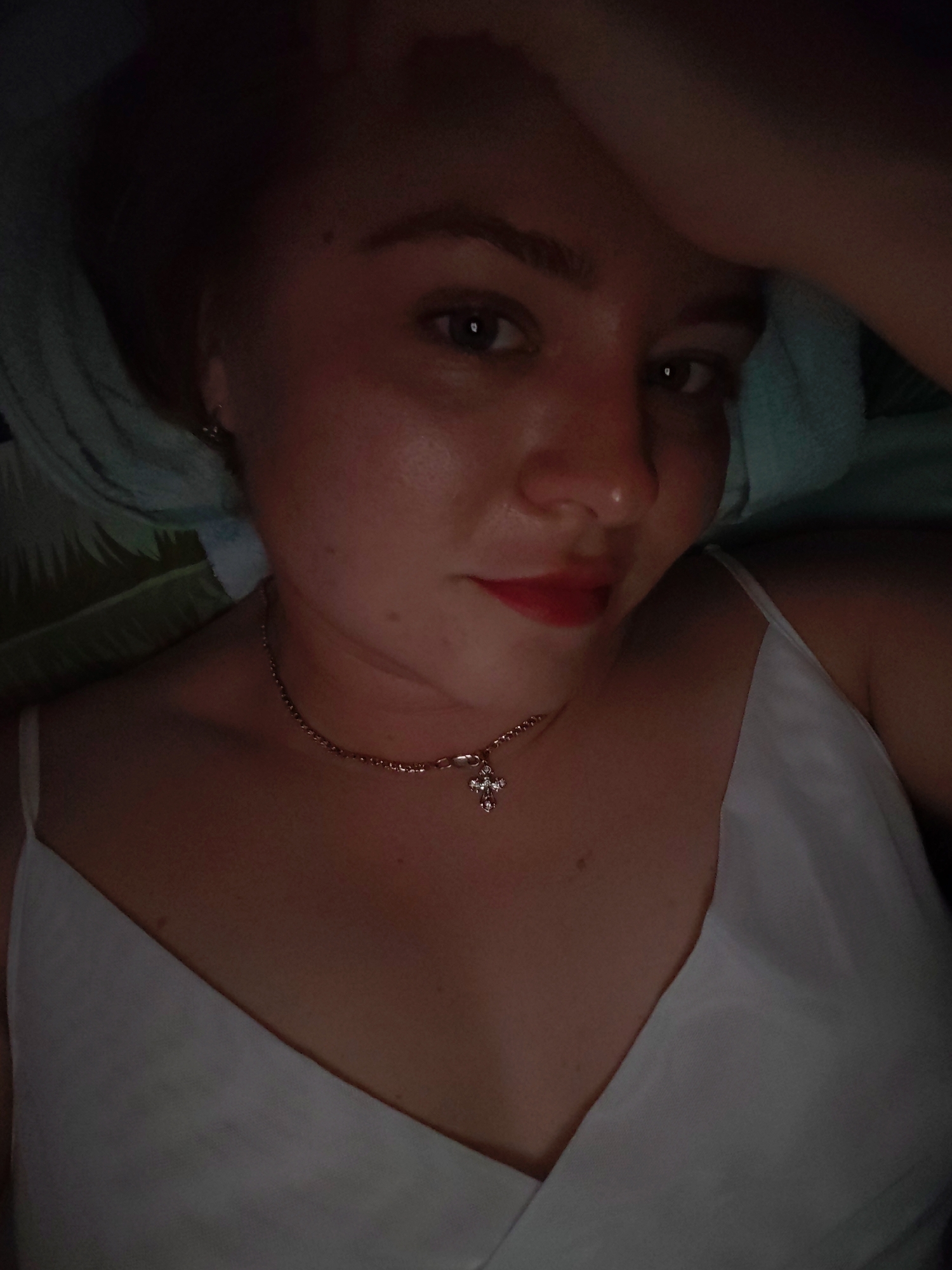 A woman with light skin, reddish hair, and red lipstick is lying down. She is wearing a white top with spaghetti straps and a necklace with a cross pendant. Her right arm is raised and her hand is near her head. A light-colored fabric is visible behind her head. The image is somewhat dark, with the light source being unclear. The location in the city of Izhevsk, Russia is not discernable from the image.