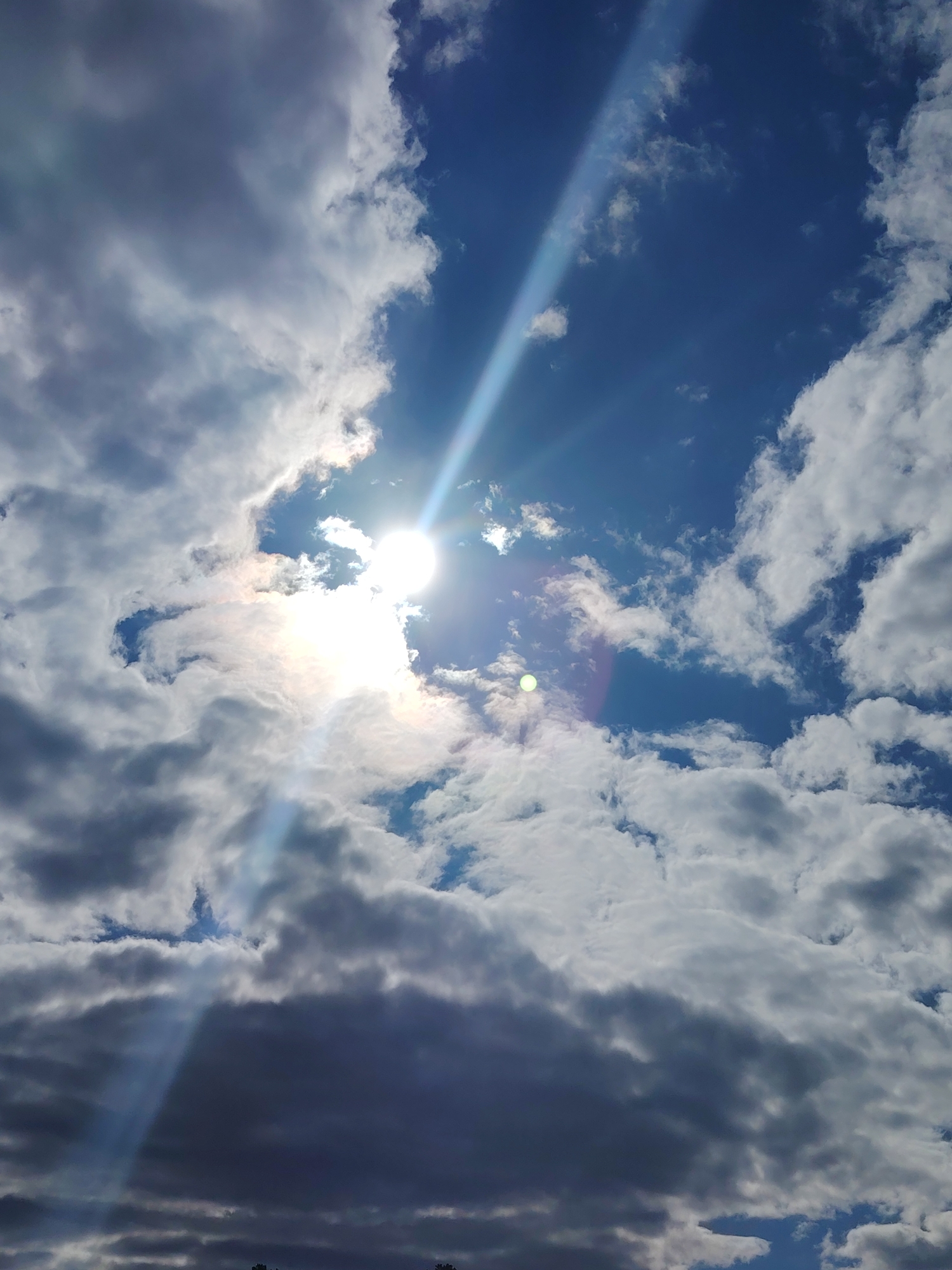 The image captures a daytime view of a partly cloudy sky. Bright sunlight streams through a gap in the clouds, creating a strong light ray. The clouds vary in color from bright white to dark gray, suggesting varying thicknesses. The sky is blue in the clearer areas. The scene is likely outdoors. The location in the city of Noviy, Russia is not discernible from the image. There are no people, subjects, objects, actions, or interactions visible. The only objects that can be seen are the clouds and the sun.