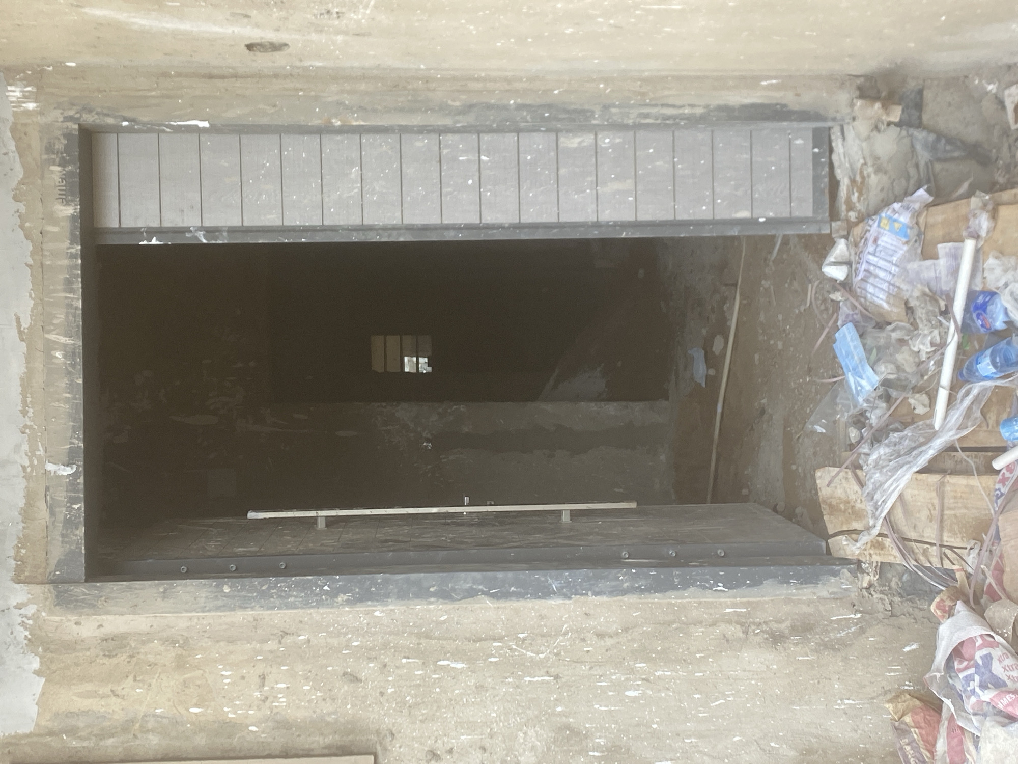 The image presents a downward perspective into a deep, dark rectangular opening, indicative of an unfinished basement, an excavated pit, or an access point to a lower level in Gosa, Nigeria. The immediate foreground consists of rough, unrefined concrete or dirt ground, speckled with white, possibly paint or plaster residue.
The entrance is framed by a sturdy, dark-colored structure, likely metal or treated wood. Across the top of this opening, a horizontal section is visible, made up of light-grey, rectangular panels or slats, which could function as a vent, a partial cover, or an architectural detail. Faint, partial text, possibly "EMENT," is discernible on the left side of this upper frame.
The interior of the opening is predominantly dark, suggesting significant depth or absence of light. Far below, a small, gridded rectangular shape, resembling a window or another opening, is dimly visible against a dark background. Closer to the viewer but still within the dark void, another horizontal structure with light-colored slats, similar to the one at the top, forms what appears to be a floor or a temporary platform. A thin, light-colored bar, supported by small vertical posts, spans this lower section.
To the right of the main opening, a considerable pile of construction debris and waste is accumulated. This includes crumpled plastic bags, several blue plastic water bottles (some empty, some with contents), sections of white PVC pipes, and pieces of cardboard or wood. Some of the discarded bags resemble empty cement sacks, with partial text such as "xtra" and "BASIC" visible on them.
No people are present, and there is no apparent activity taking place. The scene suggests a paused or ongoing construction project, characterized by raw materials and discarded waste, typical of a developing site. The ambient daylight illuminates the entrance, creating a stark contrast with the unlit depths below.