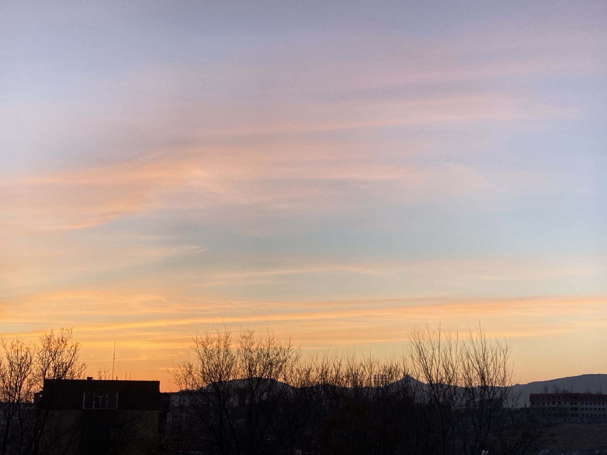 The image captures a cityscape at dusk. The sky transitions from orange near the horizon to pastel pink and light blue above. Thin, horizontal cloud formations streak across the sky. Silhouetted against the vibrant sky are trees and buildings. A multi-story building with a flat roof is visible on the left. A darker mass of trees obscures much of the mid-ground. Further in the distance, a range of hills is visible along with another building on the right side of the frame. The location appears to be Zhongtun, China.