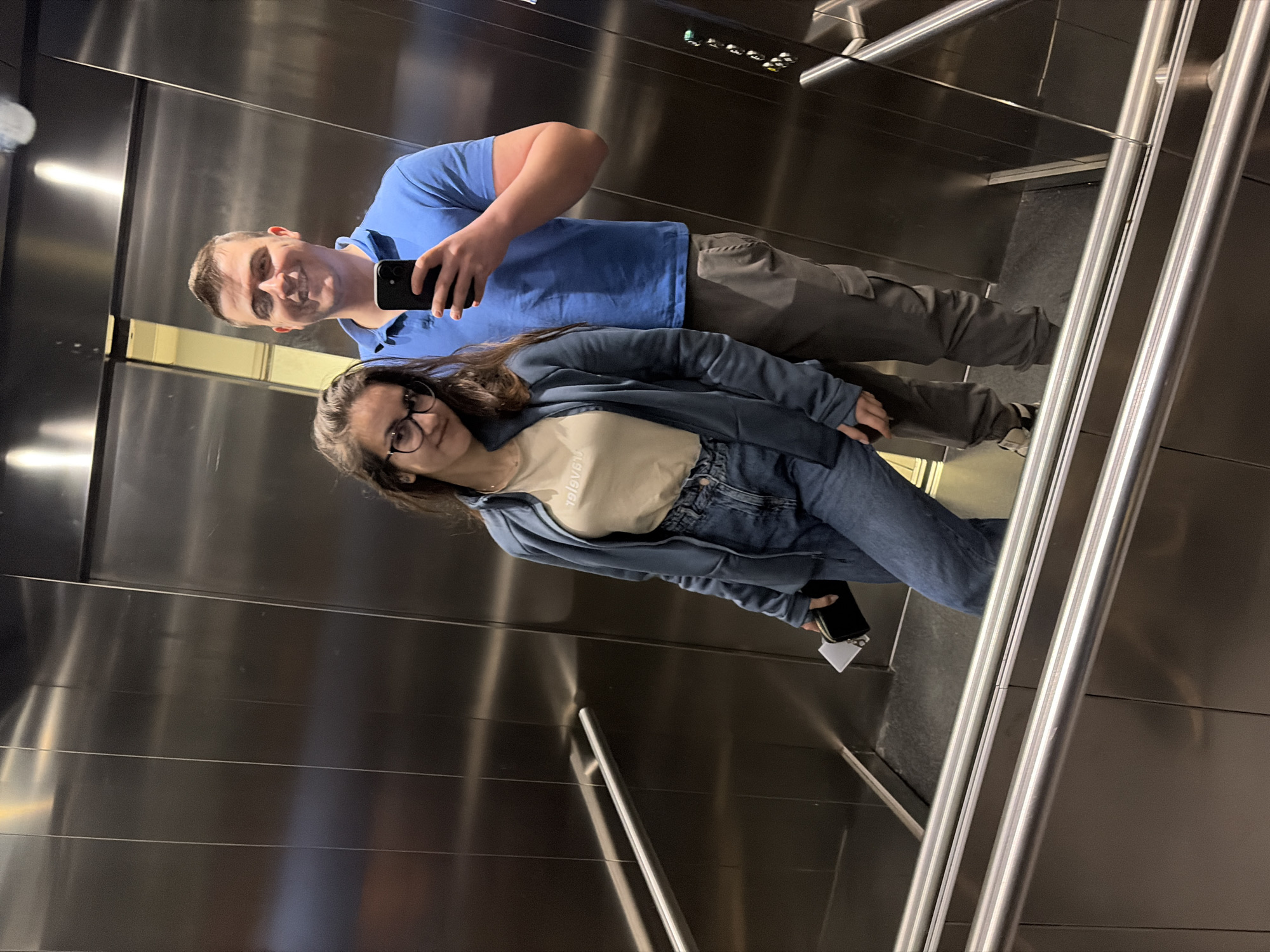 Two individuals are taking a selfie in an elevator with stainless steel walls and handrails. The man is wearing a blue polo shirt and gray pants and is holding a smartphone. The woman is wearing glasses, a light-colored t-shirt, a blue denim jacket, and jeans. The elevator appears to be in a modern building. I am unable to determine the location in the city of Sofia, Bulgaria.