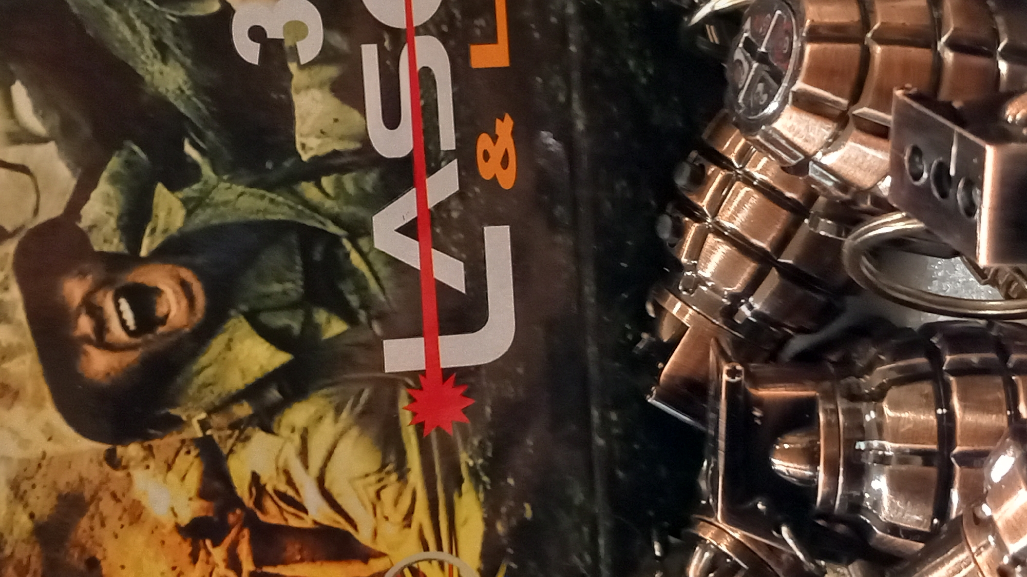 The image displays a close-up of a promotional item, likely a keychain or collectible, featuring miniature grenades. The background is a printed graphic of a soldier shouting during combat, with the words "LASER" and "& L" visible. The lighting suggests an indoor environment, possibly a retail display. The specific location in the city of Kos, Greece, cannot be determined from the image alone.