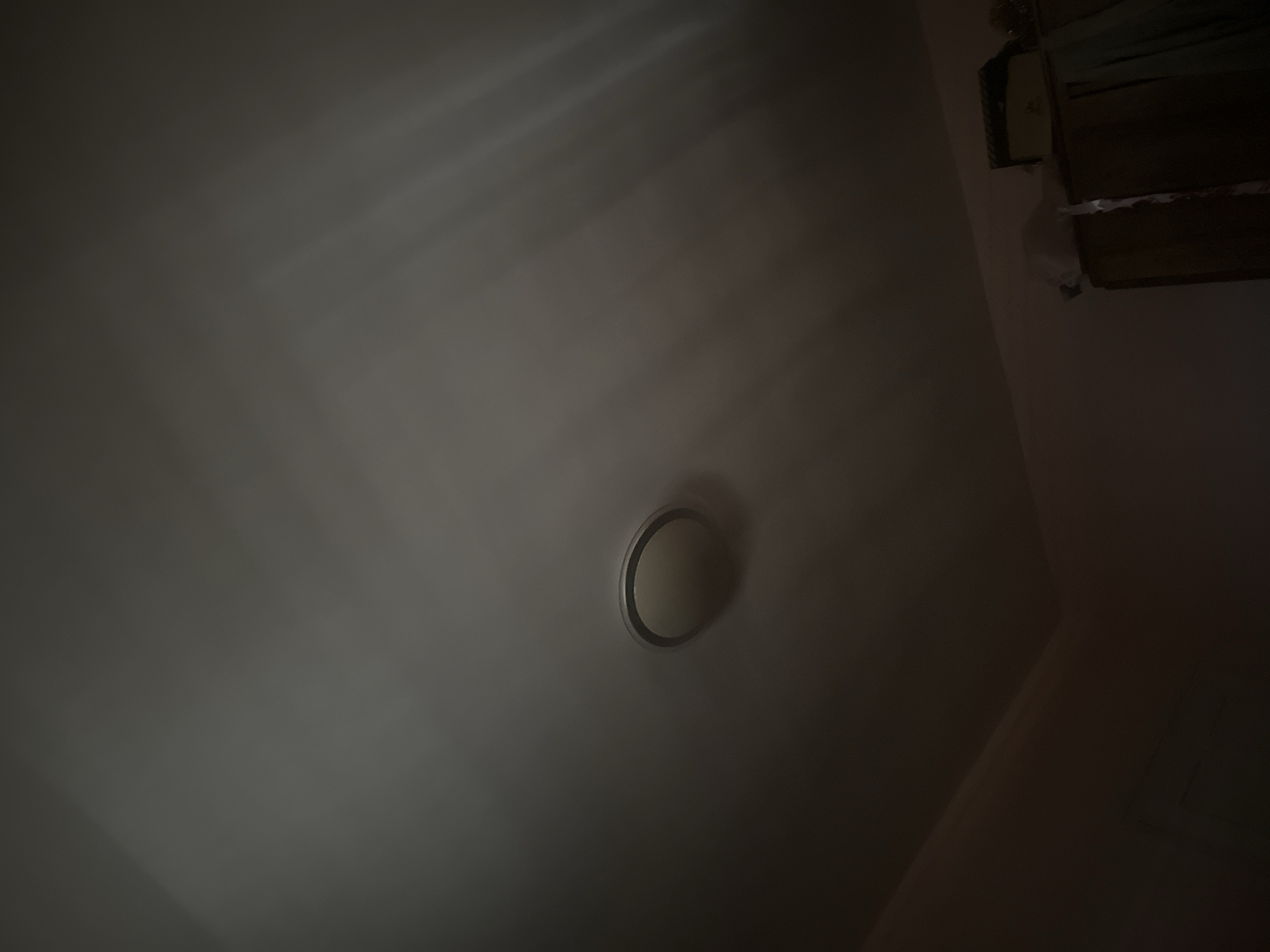 The image shows a dimly lit indoor space, likely a room, featuring a light source emitting rays across the ceiling. A circular vent is visible on the ceiling, positioned toward the lower center of the frame. In the upper right corner, there appears to be damaged or unfinished construction, possibly involving wood or plaster. The overall impression is one of poor lighting and possible disrepair. The location and specific details about Razhevo Konare, Bulgaria, cannot be determined from the image alone.