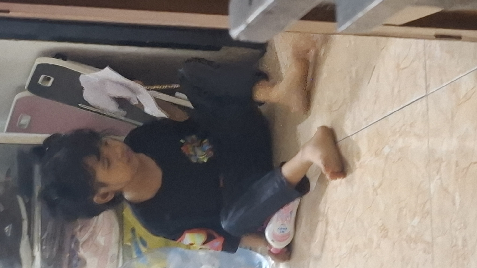 The image shows a young person with dark hair, wearing a dark shirt, sitting on a tiled floor. The person is looking downwards, with their head tilted slightly. They are wearing dark pants. The scene appears to be indoors, possibly a room or hallway, with a wooden structure visible in the background. There are several objects on the floor near the person, including what looks like a pink phone case, a white cloth, and a baby powder bottle. The tiled floor is a light beige color with darker grout lines. A thin wire or string runs across the floor. The lighting suggests it might be daytime, but the specific time of day is not clear. The overall impression is a casual, everyday scene. There is text on the baby powder bottle which appears to say "BABY". The location context is Cibinong Hilir, Indonesia.