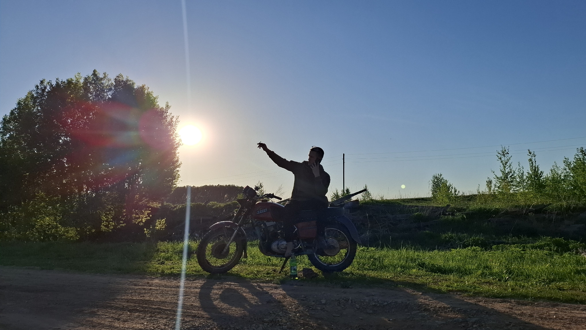 A man sits on a red motorcycle on a dirt road near Шешурга, Russia. The motorcycle is parked on a kickstand in a grassy area. A large tree is to the left of the motorcycle, obscuring the setting sun, creating a lens flare effect. The man is silhouetted against the bright sky, and is on the phone while raising his other hand. Power lines stretch across the sky in the background. Bushes and trees line the horizon.