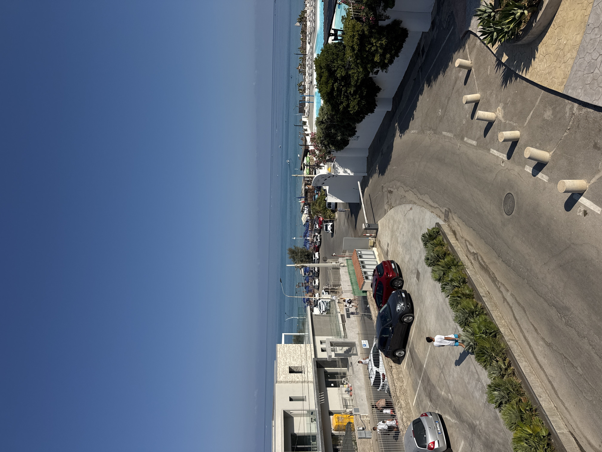 An elevated, wide-angle, eye-level shot captures a sun-drenched scene in Capaci, Italy, with the Tyrrhenian Sea dominating the upper half of the frame. The clear, deep blue sky meets the tranquil, turquoise ocean at a sharp horizon. Scattered on the water are small boats.
In the middle ground, a resort-like setting unfolds. A large, bright blue swimming pool is visible, surrounded by numerous white lounge chairs. Beyond the pool, a beach area is set up with rows of blue umbrellas. A white stucco wall lines the perimeter of the resort, separating it from the road and parking area.
The foreground and mid-ground showcase a parking lot and the immediate vicinity of a building. A modern, three-story building with white walls and dark-trimmed windows is on the left. Adjacent to it is a smaller, gray structure with an entrance. Several cars are parked in the lot, including a silver hatchback, a dark grey hatchback, and a red SUV. A few people are visible in the parking area and walking near the building. One person in a white t-shirt and blue shorts walks across the parking lot, casting a long shadow. Another group of people, including adults and children, are walking near a metal fence.
The road leading into the parking area has a white gate. Several concrete bollards are spaced along the edge of the road. The environment appears to be a typical coastal tourist destination, bustling with activity under a clear, bright sky.