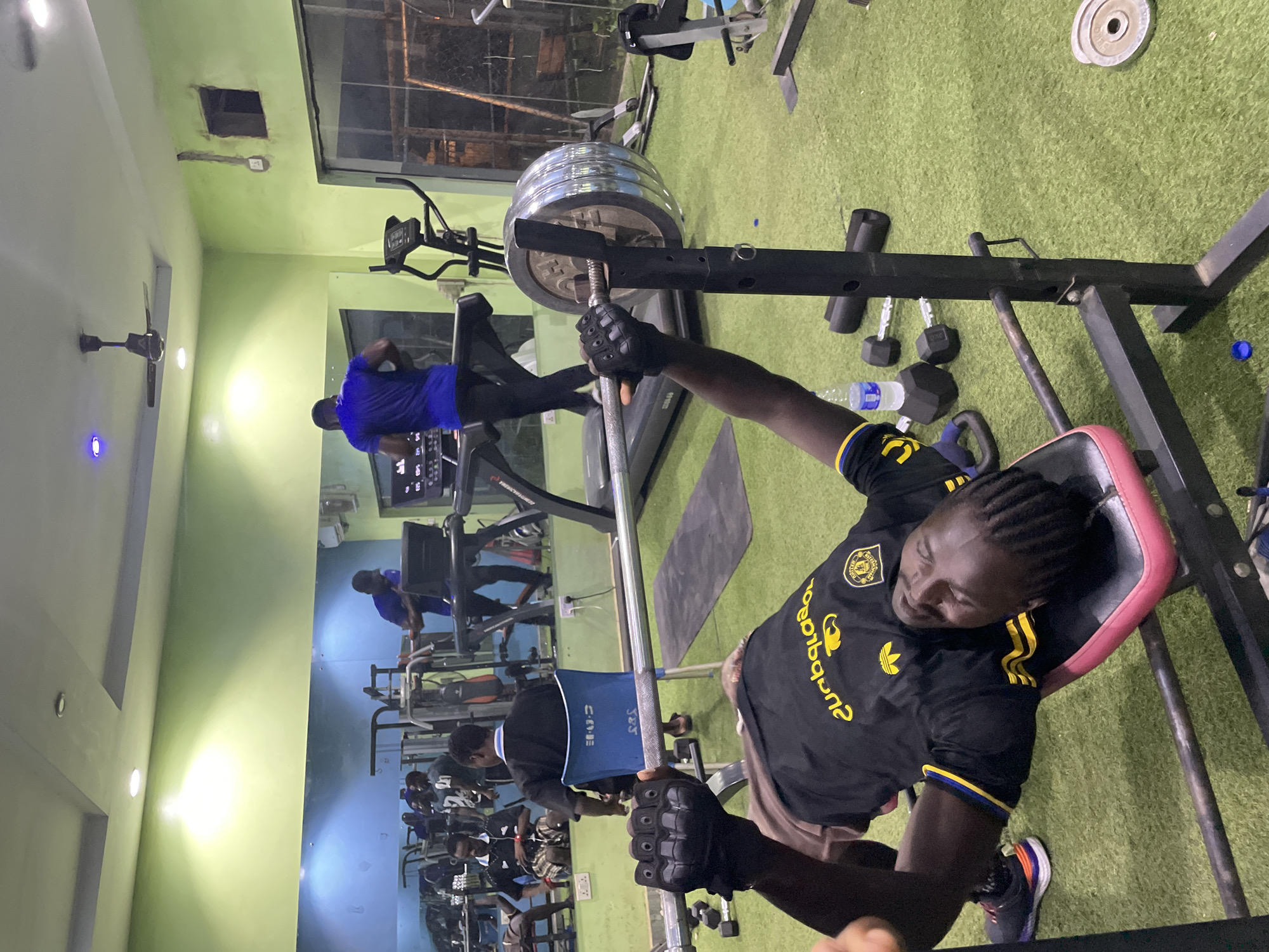 This image captures a vibrant scene inside a gym or fitness center in Akwanga, Nigeria, likely in the evening given the dark exterior visible through the windows. The walls are painted a light green, and the floor is covered with artificial green turf.
In the foreground, a person with dark skin and braided hair is lying on a pink and black weight bench, preparing for a bench press exercise. They are wearing a black short-sleeved t-shirt with yellow stripes on the sleeves and a yellow Adidas logo, as well as a crest-like emblem on the chest. Visible yellow text on the shirt, when read correctly, appears to be "Buqaana" or similar. The individual is wearing black fingerless gloves and dark athletic shoes with colorful soles, and is grasping a heavy barbell loaded with multiple metallic weight plates. A clear plastic water bottle with a blue cap rests on the floor nearby.
In the mid-ground and background, several other people are engaged in various fitness activities. One person in a blue t-shirt and black pants is seen using a treadmill. Other individuals are partially visible, reflected in a large mirror, interacting with different gym equipment. A black ceiling fan is mounted on the wall, and several bright, circular lights illuminate the space. Dumbbells and other exercise machines are scattered throughout the gym floor.