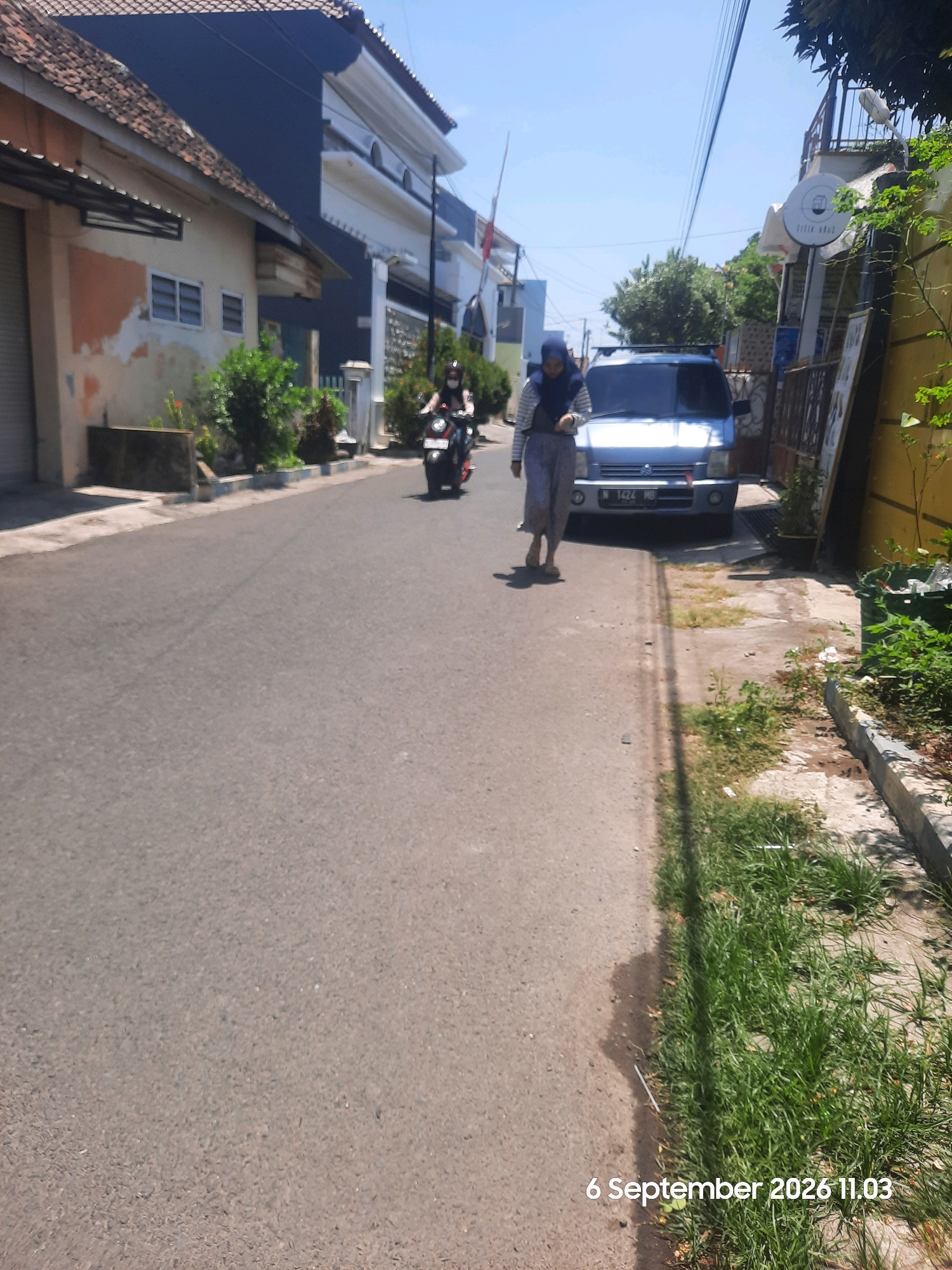 The image shows a street scene in Probolinggo, Indonesia. A woman wearing a blue hijab and a light-colored striped shirt walks on the right side of the road, carrying a small white bag. Beside her, a light blue car is parked. Further down the road, a person on a motorcycle, also wearing a hijab, is riding away from the camera. The street is paved, with buildings and vegetation lining both sides. The sky is clear and blue. The timestamp "6 September 2026 11.03" is visible in the bottom right corner.