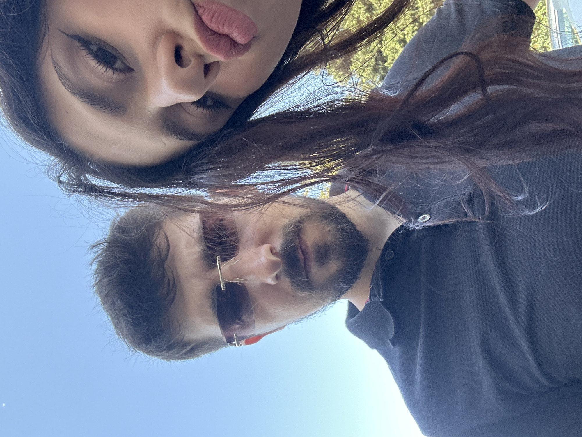 A selfie taken outdoors shows a man and a woman. The man is in the foreground, on the left side of the frame, wearing sunglasses and a black polo shirt. He has short, dark hair and a beard. The woman is on the right side of the frame, with her face close to the camera, puckering her lips. She has long, dark hair. The background is a clear blue sky and some green foliage. The image appears to be taken in a city, but there are no specific landmarks or clues that would indicate the city of Baku, Azerbaijan.
