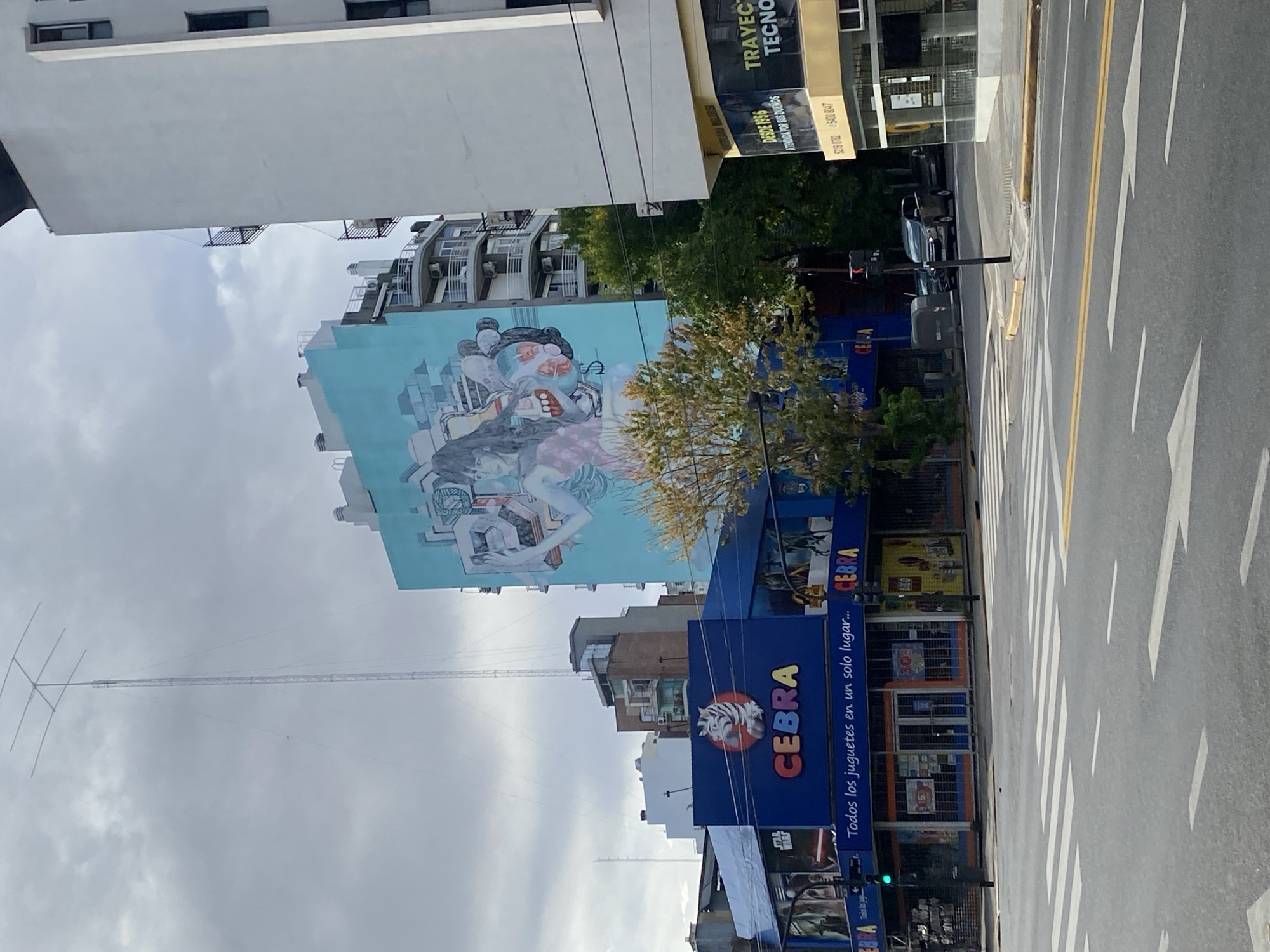 The image captures an urban scene in Buenos Aires, Argentina. A large mural depicting a woman is painted on the side of a tall building. A "CEBRA" toy store stands below the mural, its blue facade and signage visible, stating in Spanish "Todos los juguetes en un solo lugar...", followed by the company's logo. A tower in the background extends upwards into a cloudy sky. A street runs alongside the buildings, with clear markings indicating pedestrian crossings and lanes. Buildings of varying heights line the street, creating a typical cityscape. A sign "TRAYEC TECNO" is visible on another building. The image appears to be taken on a cloudy day with ambient light.