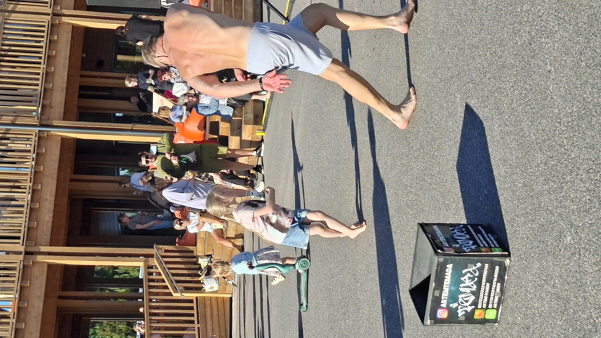 This image captures an outdoor event with a group of people gathered on a sunny day. The main subjects are several individuals, including adults and children, interacting in a casual setting. A shirtless man with tanned skin is prominently featured, appearing to be in motion or mid-activity. Two young girls are also visible; one is on a scooter, and the other is standing nearby. The scene appears to be set in a park or outdoor venue with a wooden structure in the background, possibly a building or a stage. The ground is asphalt, suggesting a paved area. Shadows are cast on the ground, indicating it is daytime, likely mid-afternoon given the angle of the sun.
A small black box with colorful branding is present in the foreground. Visible text on the box includes "ARTEMTEMADA" with an Instagram logo, "FANDR" in a stylized font, and various alphanumeric codes and names like "AKTIIVNE IVANOV" and "ARTEM TEMADOV". The overall atmosphere seems relaxed and festive. The presence of a scooter and the casual attire of the people suggest a family-friendly event or public gathering. The lighting and the presence of shadows point to a clear, sunny day. The location context provided is Tallinn, Estonia.